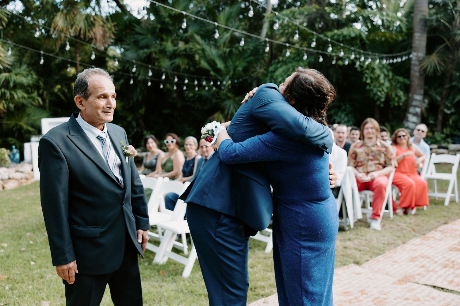 Guests at an outdoor wedding ceremony greet each other with hugs under string lights and trees.
