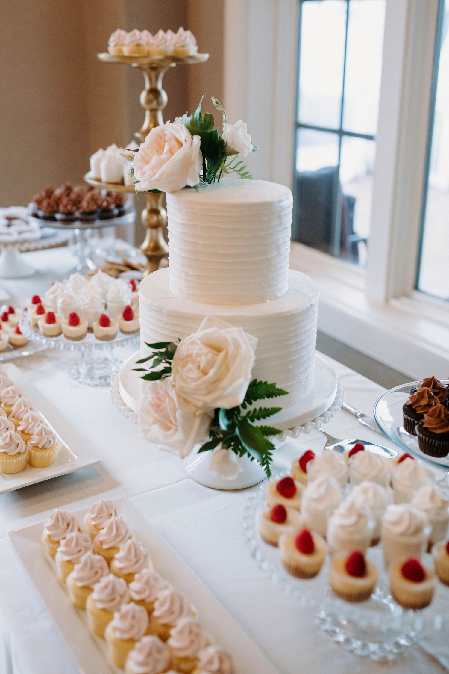 Elegant white wedding dessert table with tiered cake, cupcakes and sweet treats.