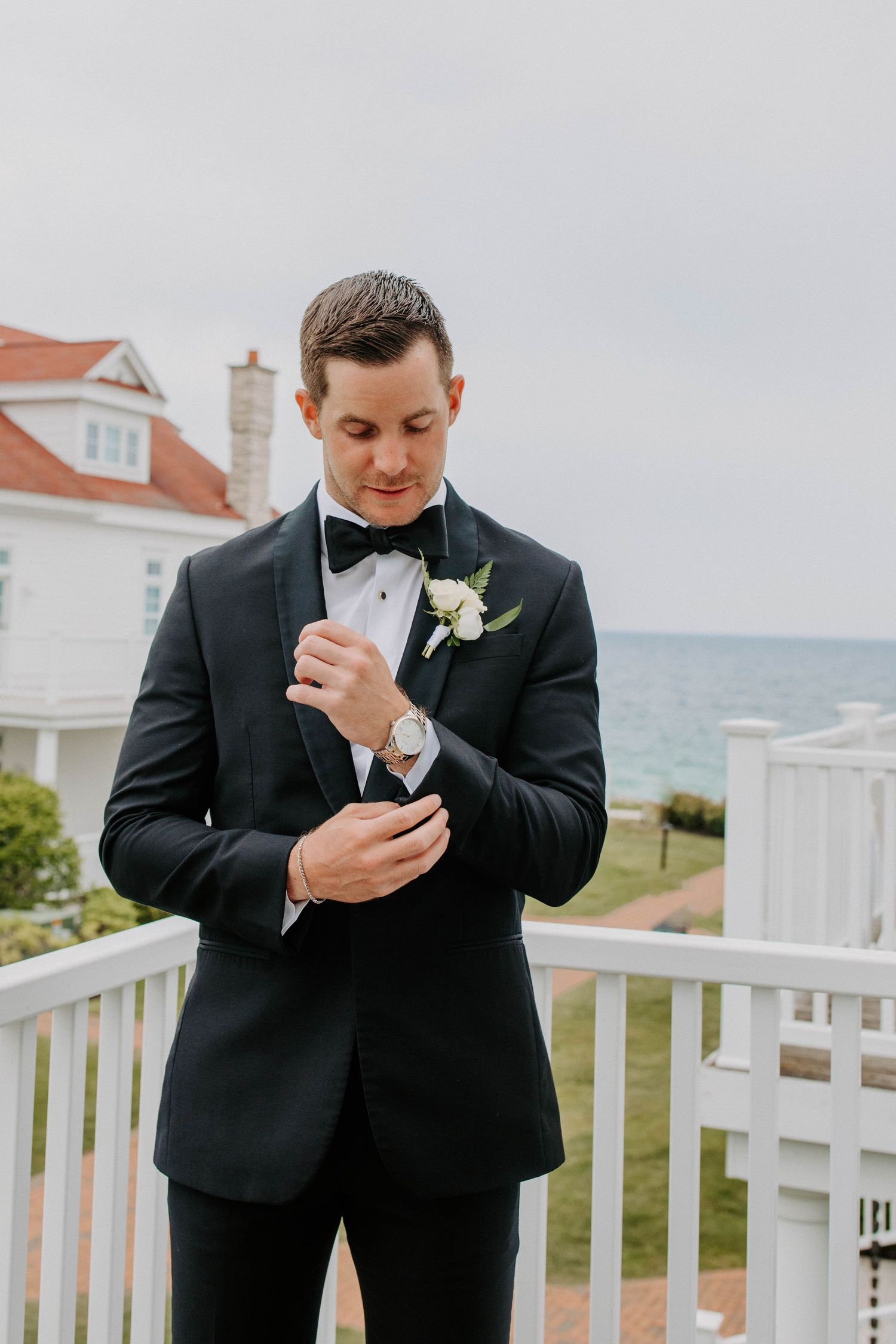Groomsman in black tuxedo adjusts his boutonniere on a white porch railing by the ocean.