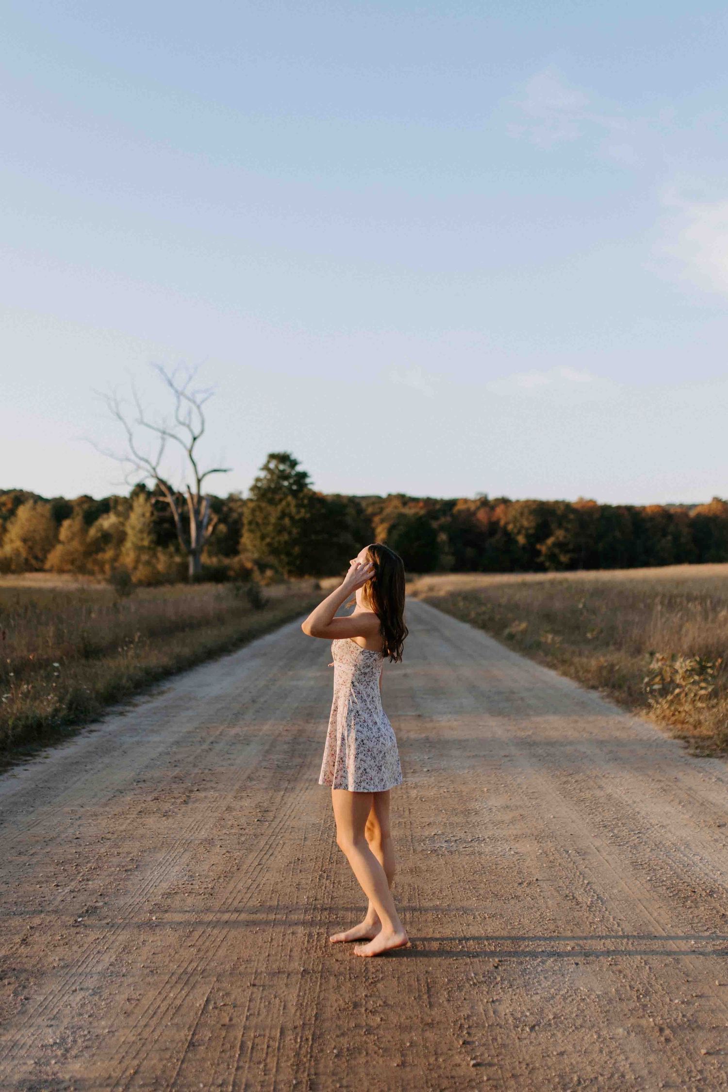 A person in a floral dress dances barefoot on a dirt country road during golden hour.