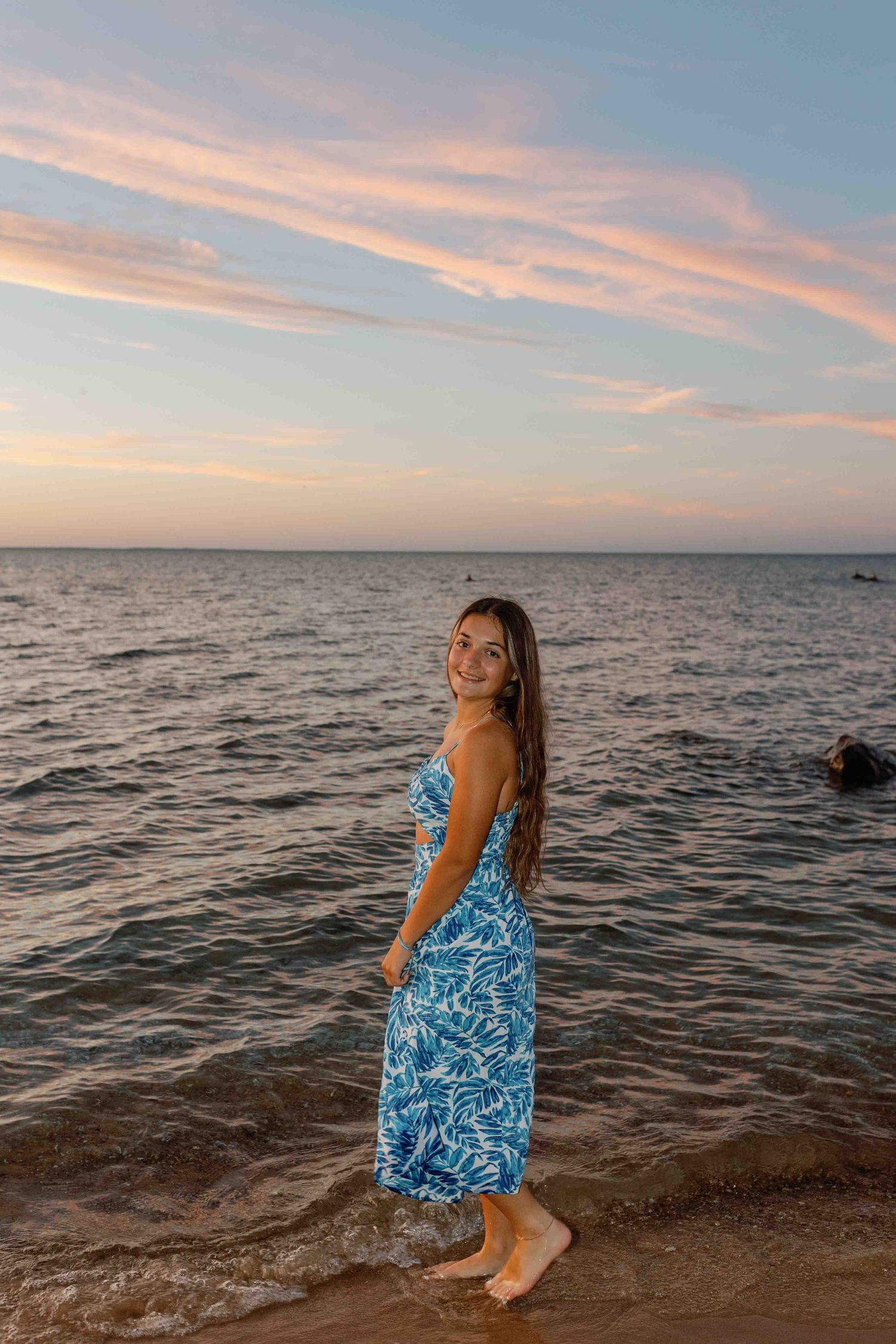 A figure in a blue patterned dress poses along a rocky beach shoreline at sunset.