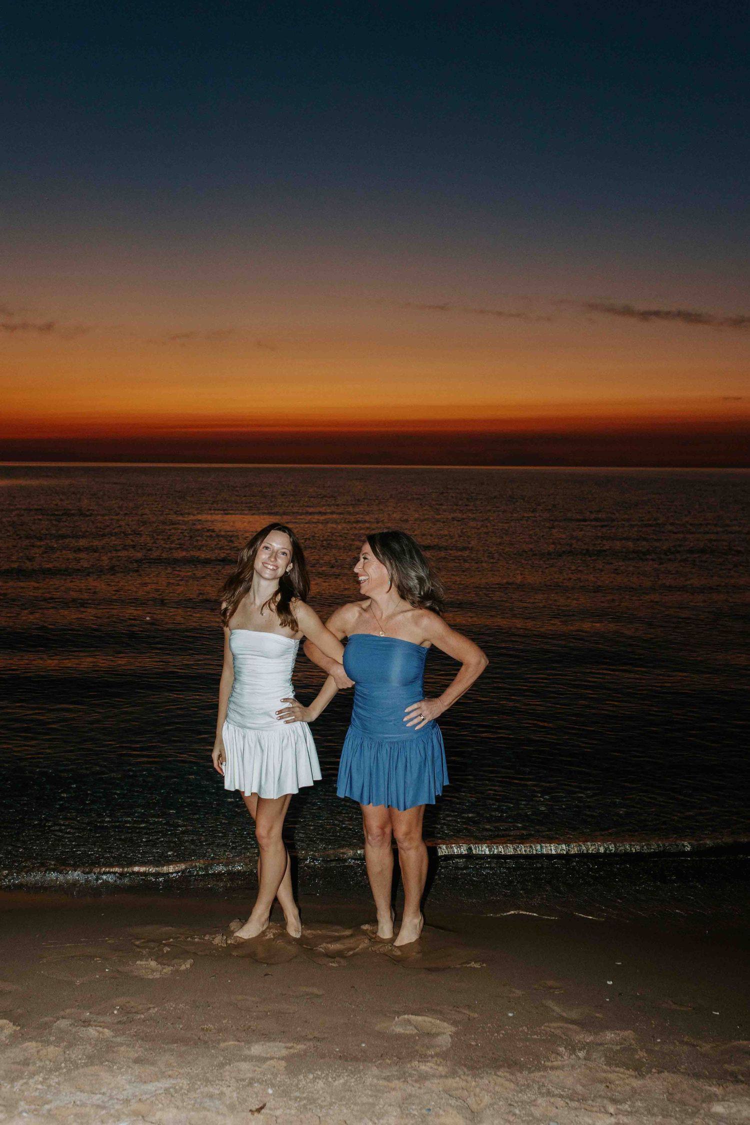 Two people strike playful poses on the beach with a stunning orange sunset behind them.