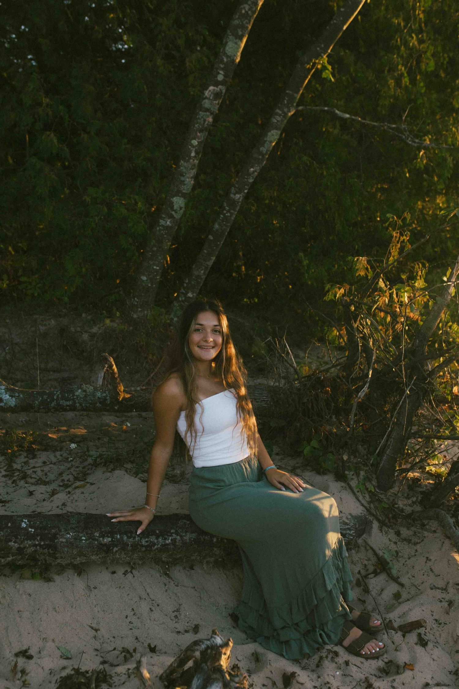 A person in summer attire poses against a sandy bank with sunlit trees in the background.