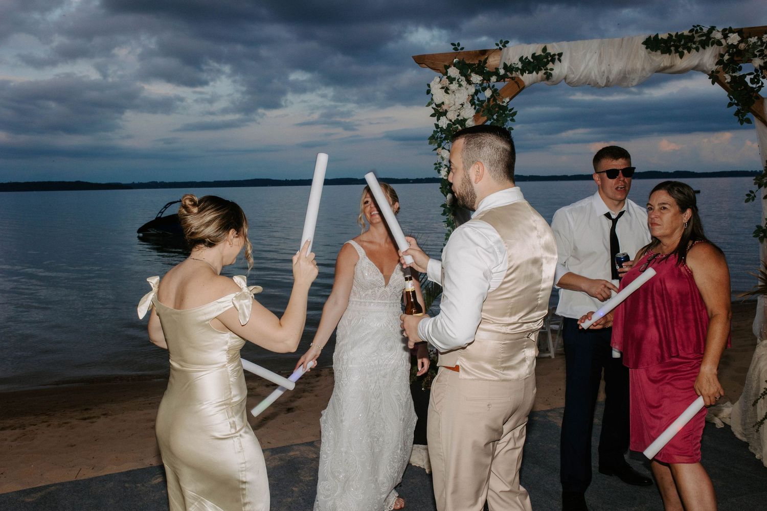 Wedding guests in formal attire hold light sabers on a beach at dusk with stormy clouds overhead.