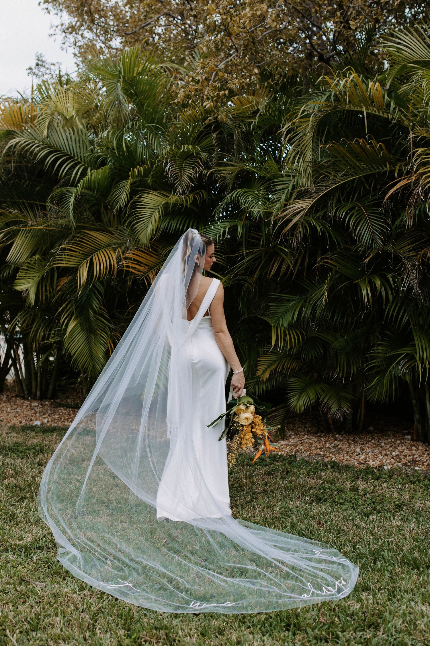 A bride in a sleek white gown with flowing veil poses against tropical palm trees.