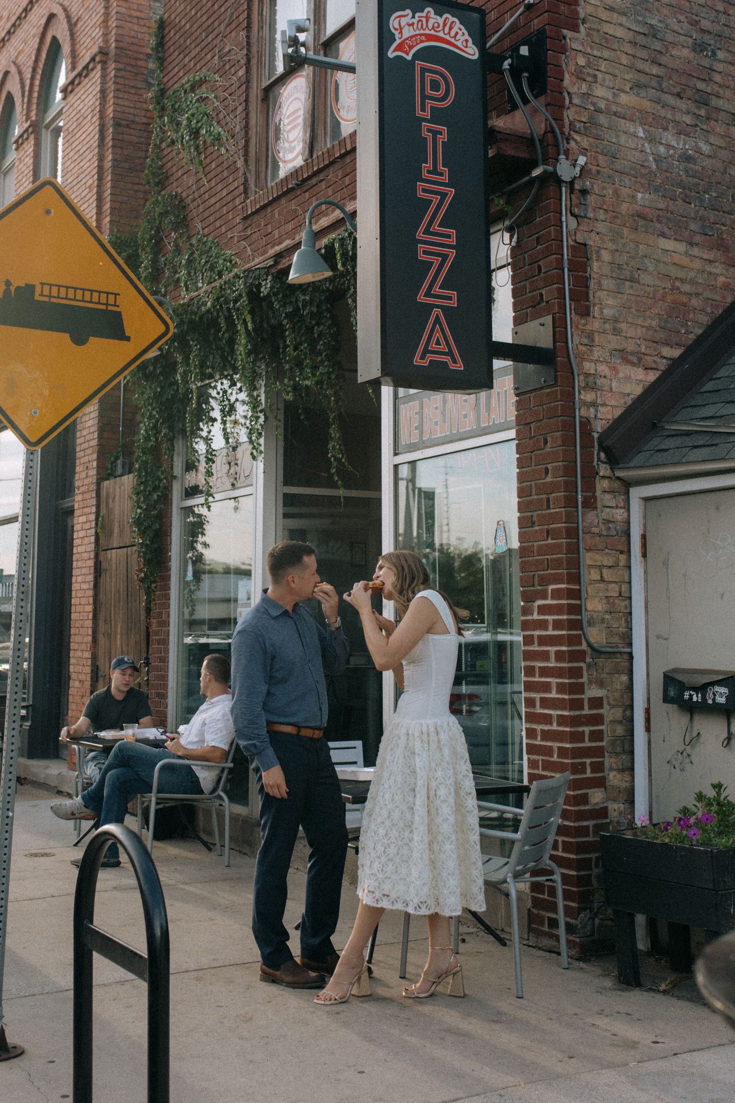 A couple stands outside a brick building with a pizza restaurant sign and storefront.