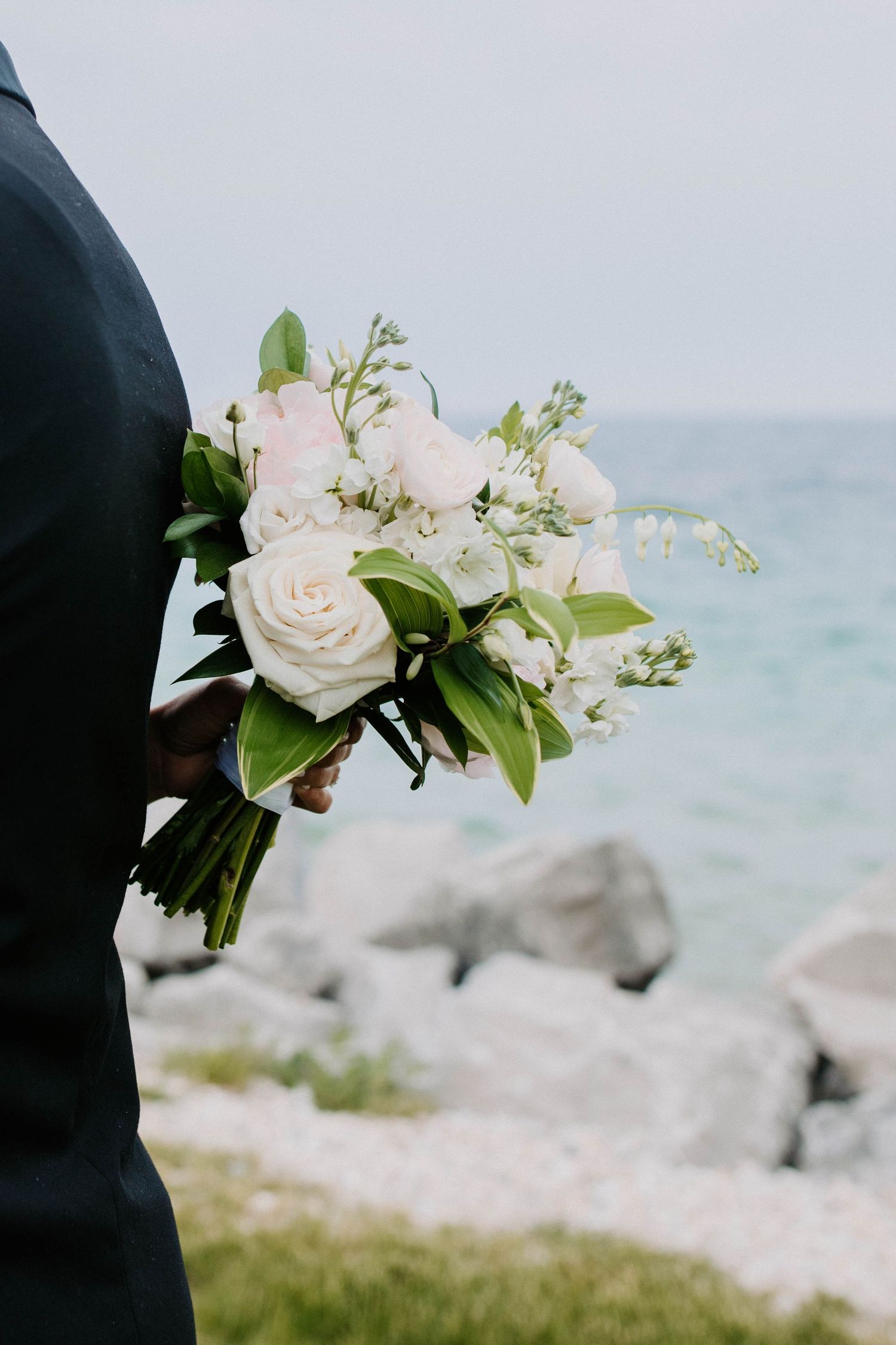 White roses and greenery in wedding bouquet against coastal background.