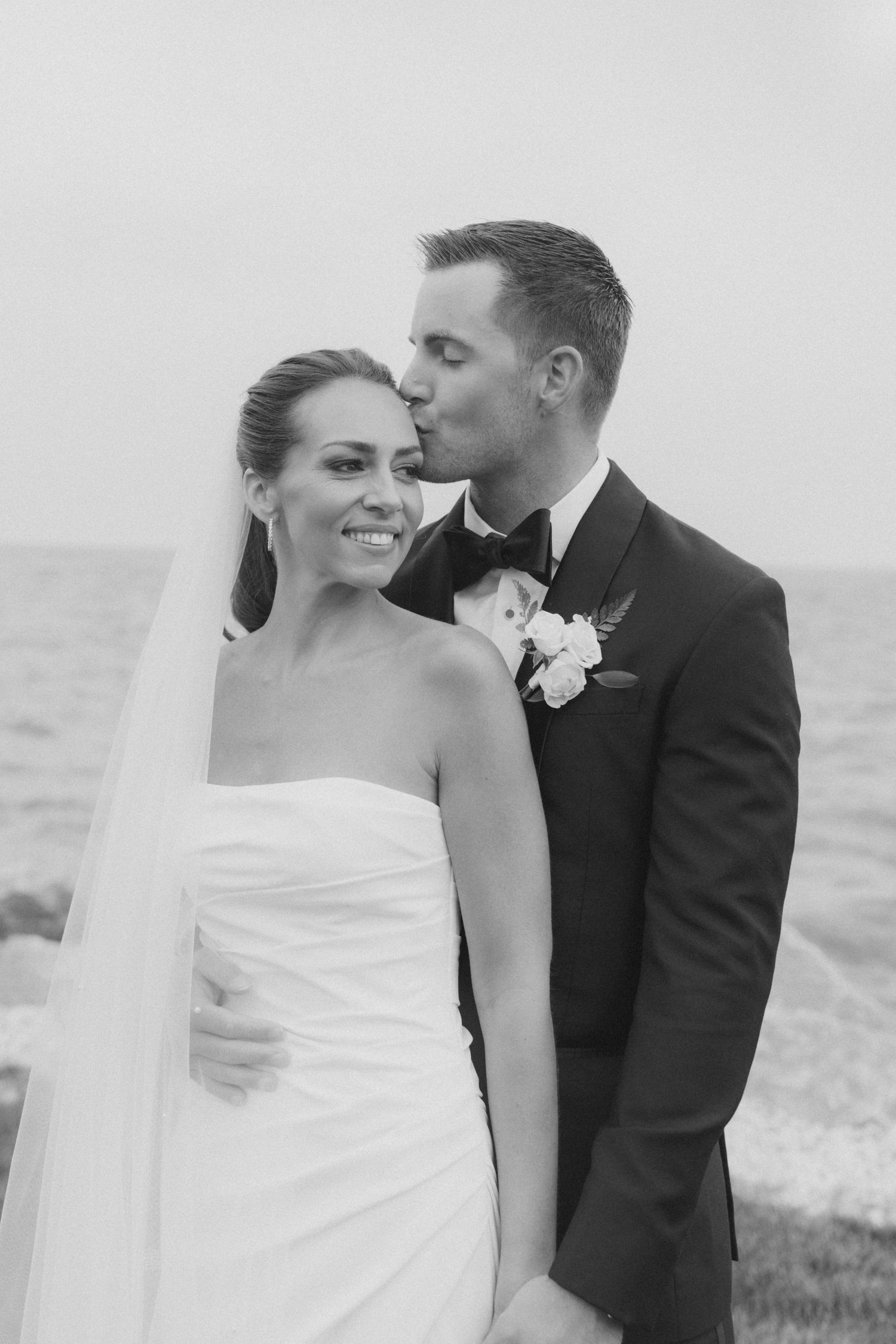 A romantic black and white wedding photo of a couple by the ocean with the groom kissing the bride's temple.