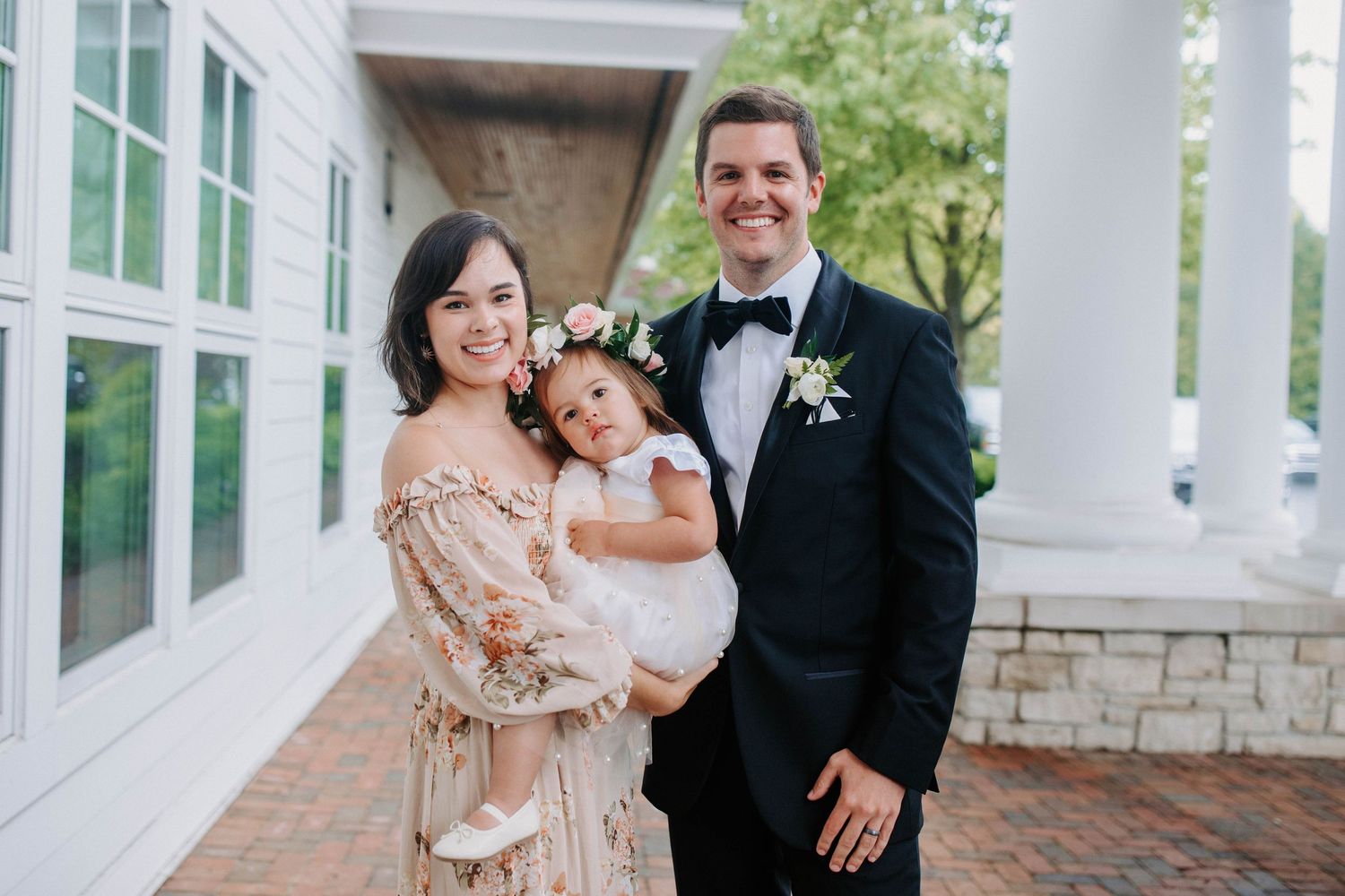 A couple and child pose together on a white columned porch during a formal event.