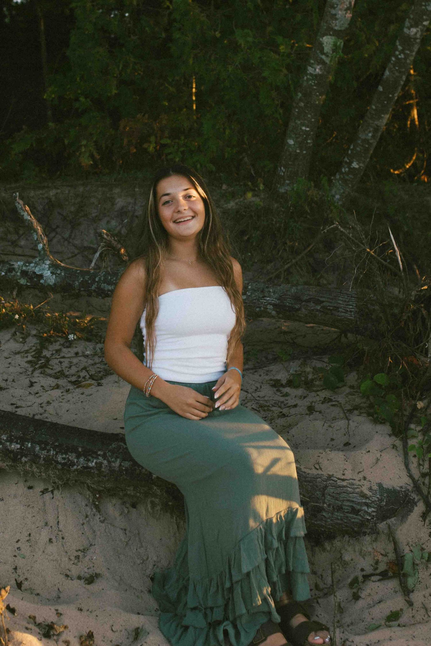 A person in a white top and green ruffled maxi skirt poses on a sandy bank against a wooded background.