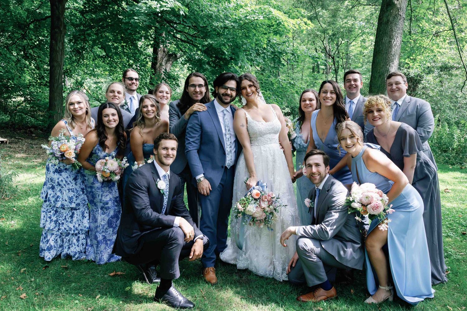 Wedding party gathered for casual group photos in a lush green outdoor setting.