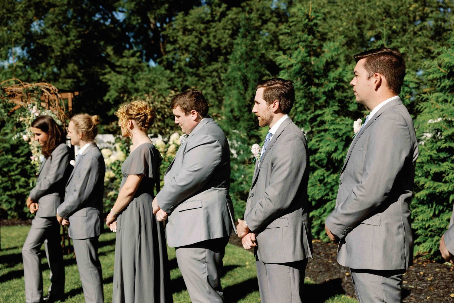 Groomsmen in matching grey suits standing in line during outdoor wedding ceremony.