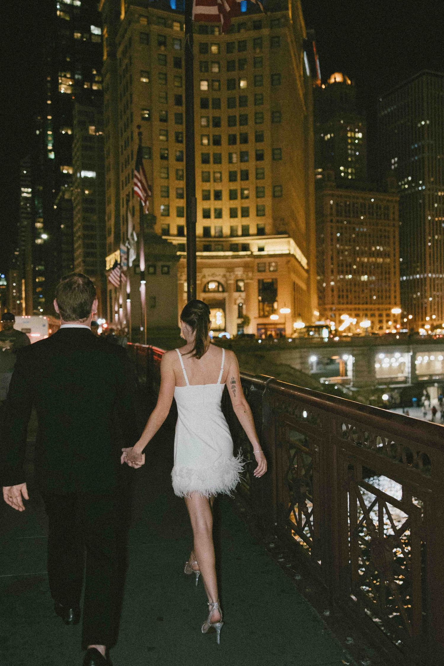 Couple walks along Chicago's riverwalk at night with illuminated buildings in the background.