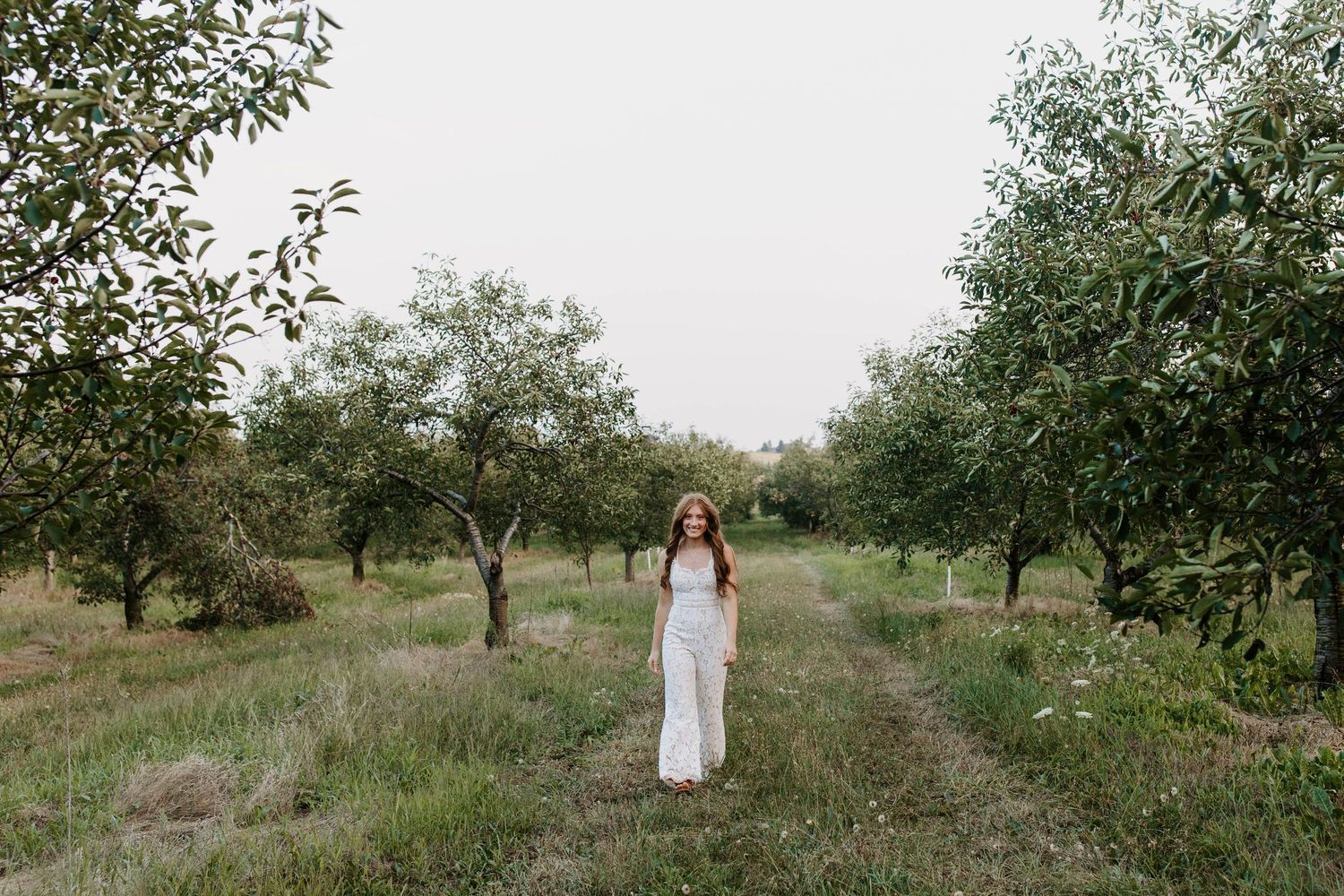 Person in white dress walks through apple orchard on sunny day.