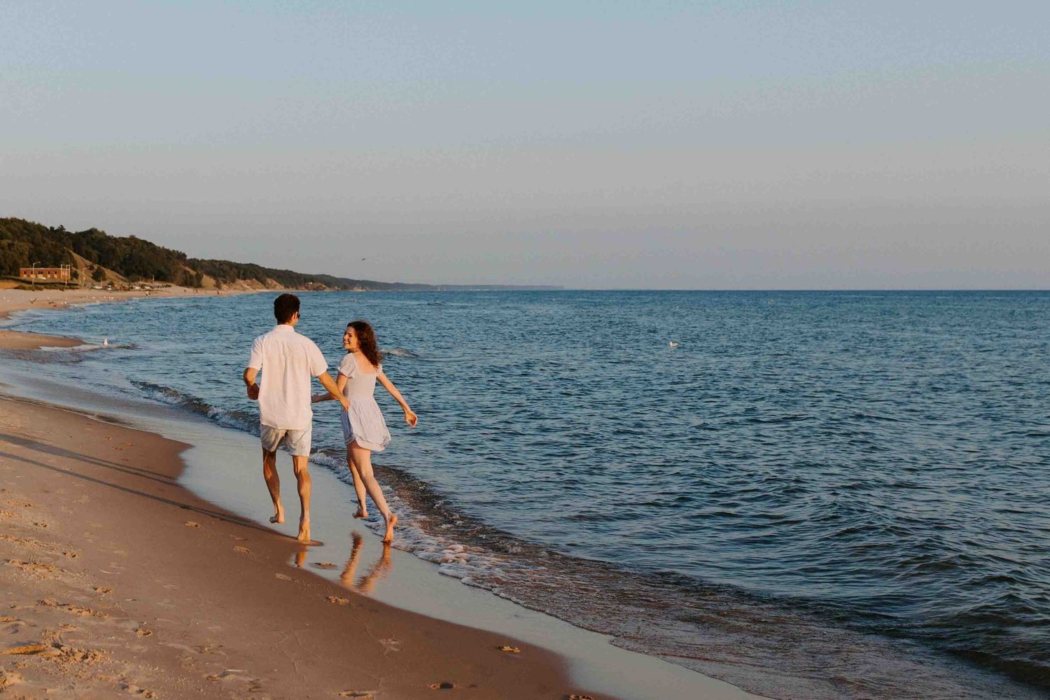 Two people stroll hand in hand on a peaceful beach with calm waters and distant hills.