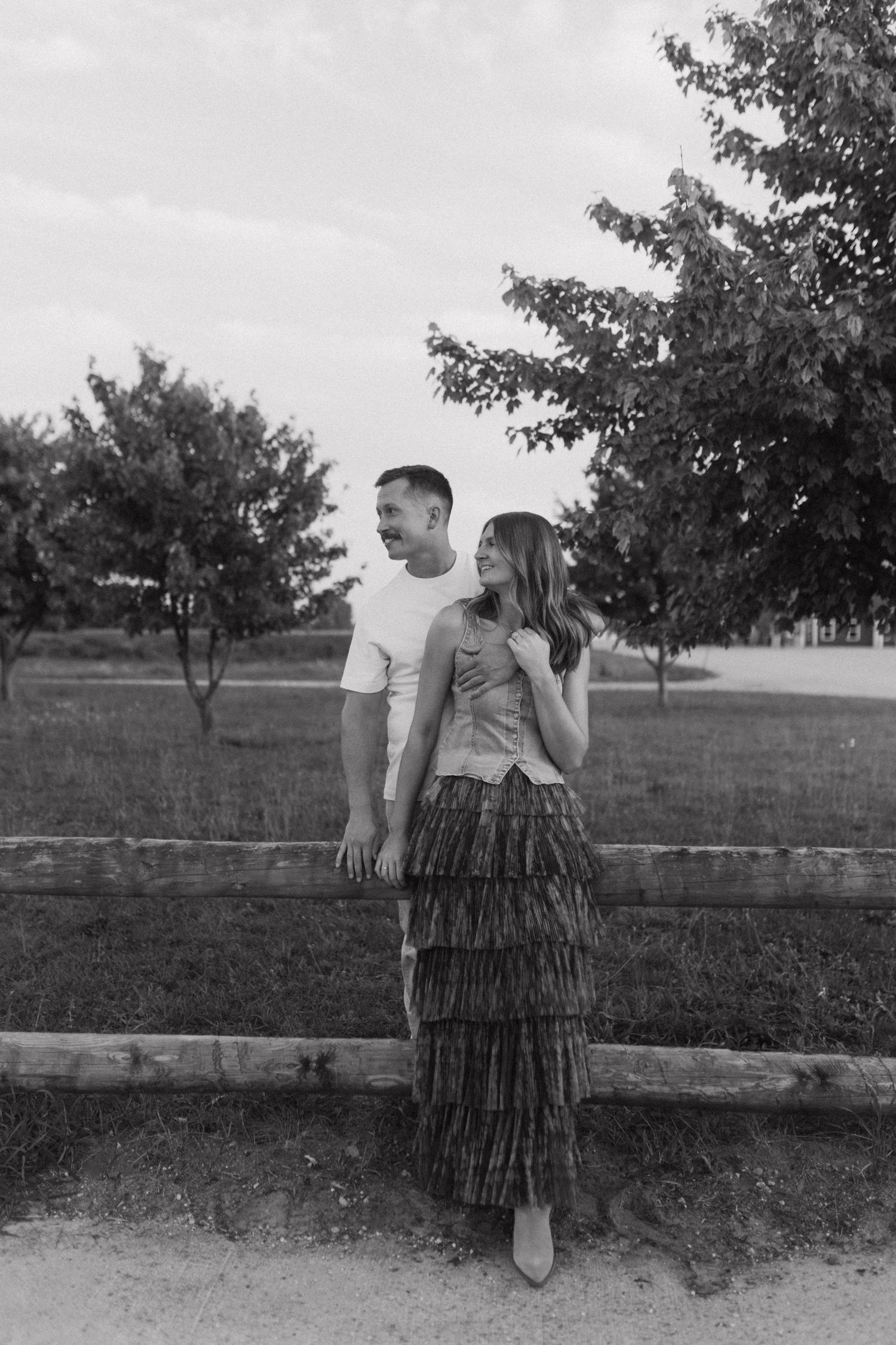 A couple sharing tender moments along a rustic fence in a pastoral setting.