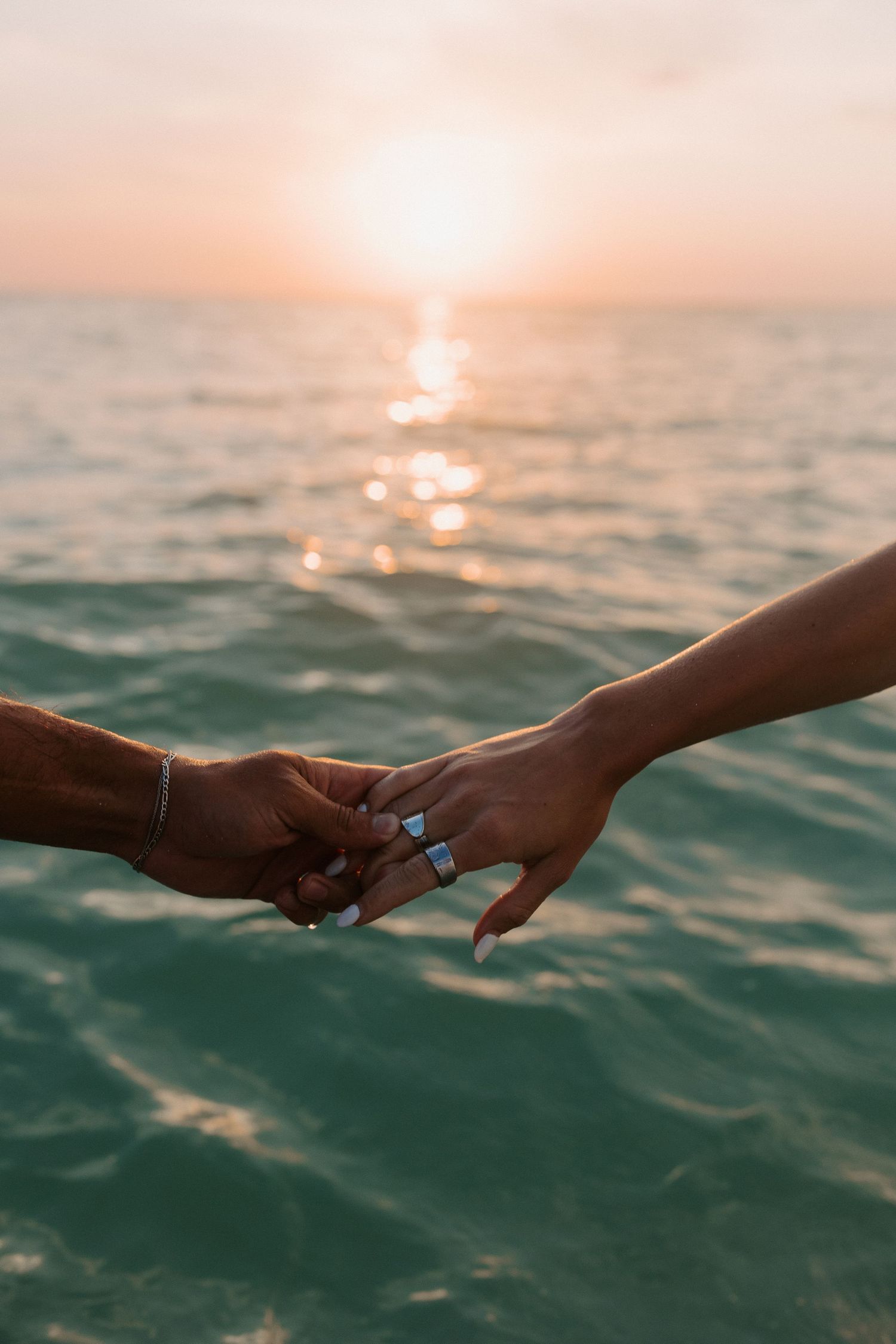 Two hands with rings reach toward each other over sunset-lit ocean waters.