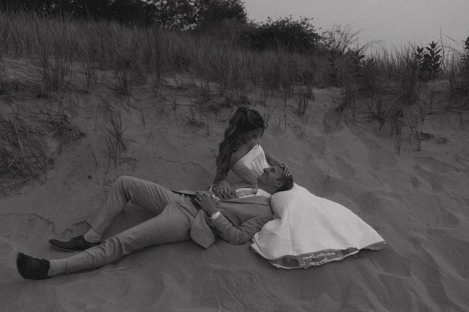 Artistic black and white beach wedding photos with couple resting on sand dunes.