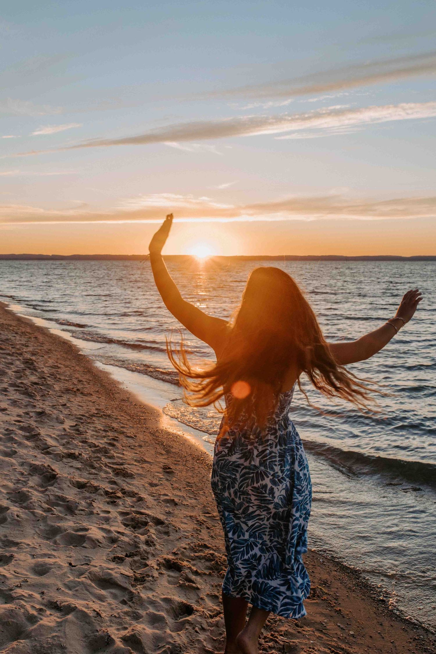 Person in patterned dress dances on beach with arms raised during sunset.
