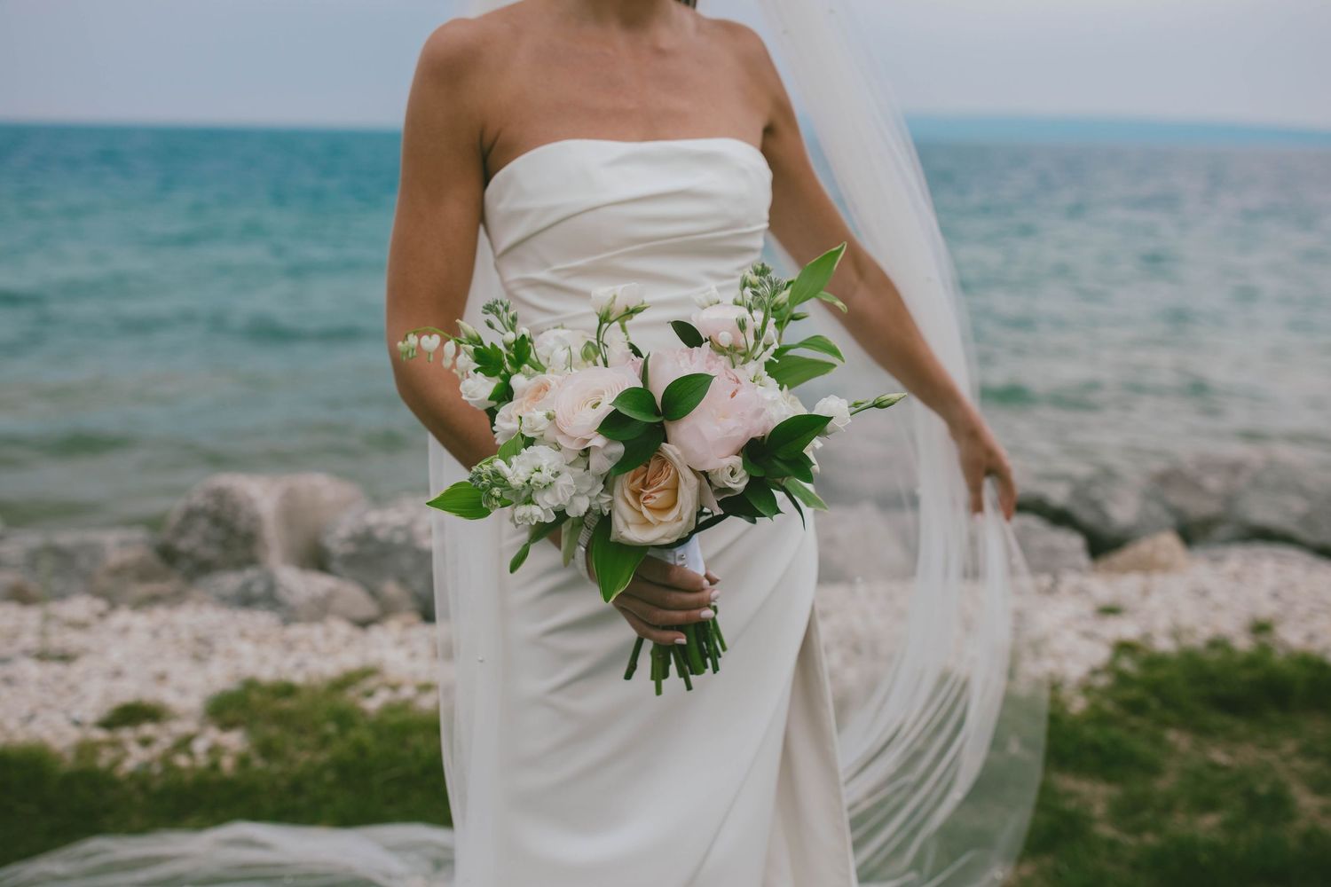 Elegant white wedding bouquet with roses and greenery against a beachfront backdrop.