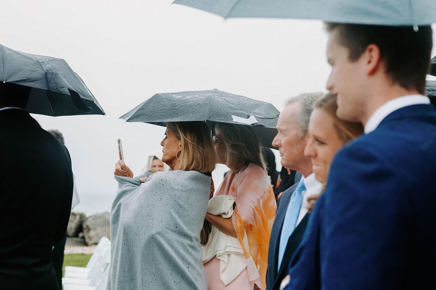 Wedding attendees stand with umbrellas during an overcast outdoor ceremony.