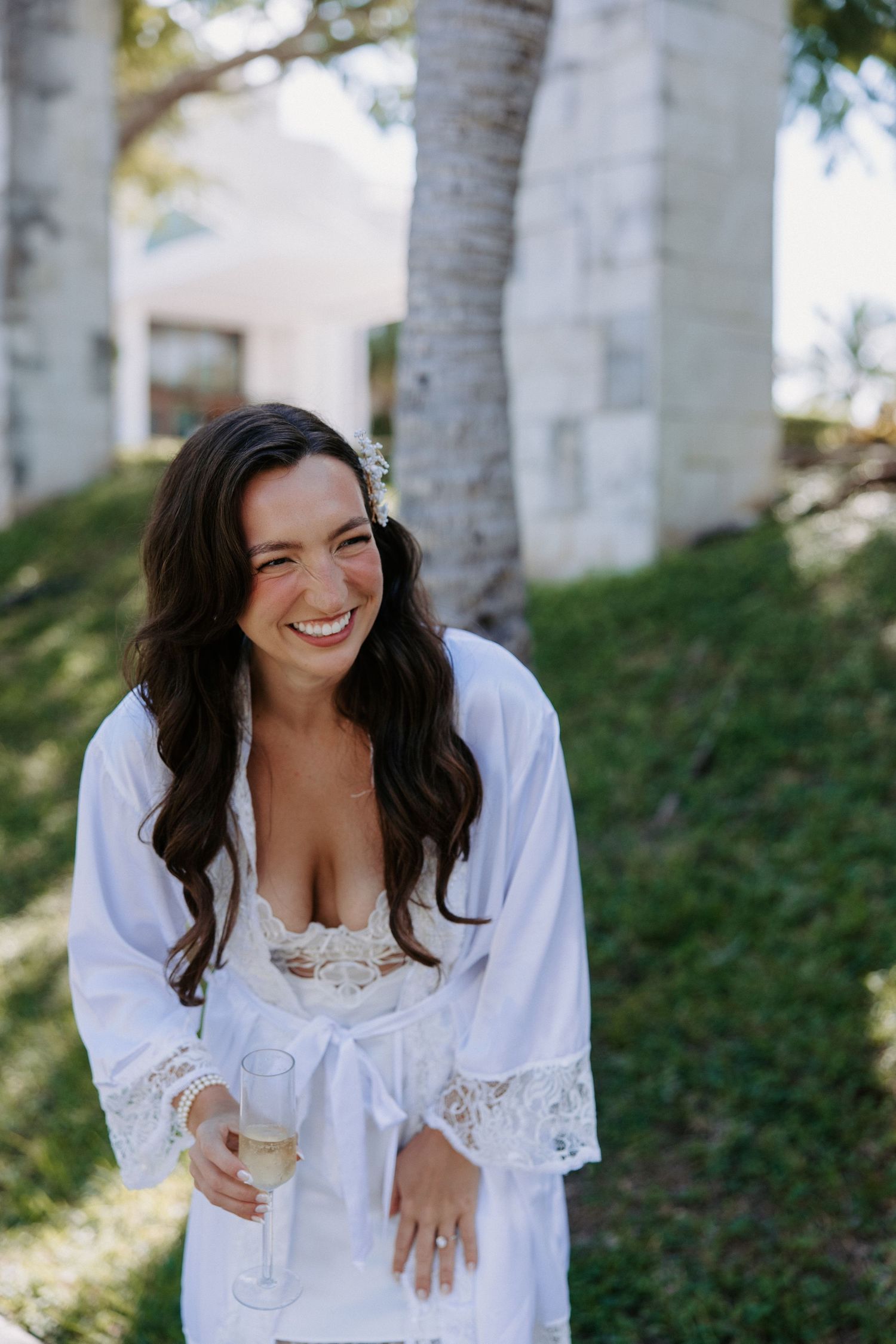 Woman in white lace robe smiling during wedding preparations outdoors.