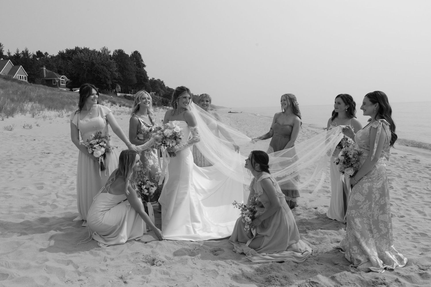 Bridesmaids in flowing blue dresses sitting on beach sand together.