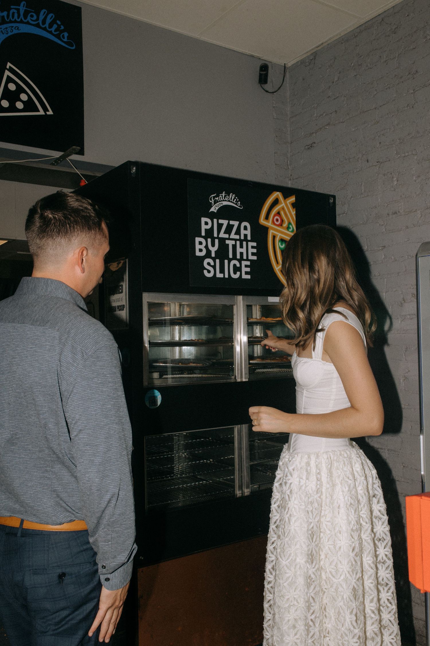 A couple being playful in front of a pizza shop with 'By the Slice' signage.