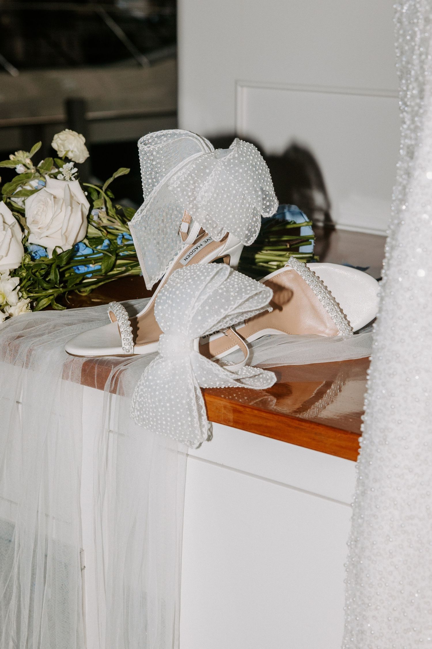 White beaded wedding shoes displayed with floral arrangements on a wooden surface.