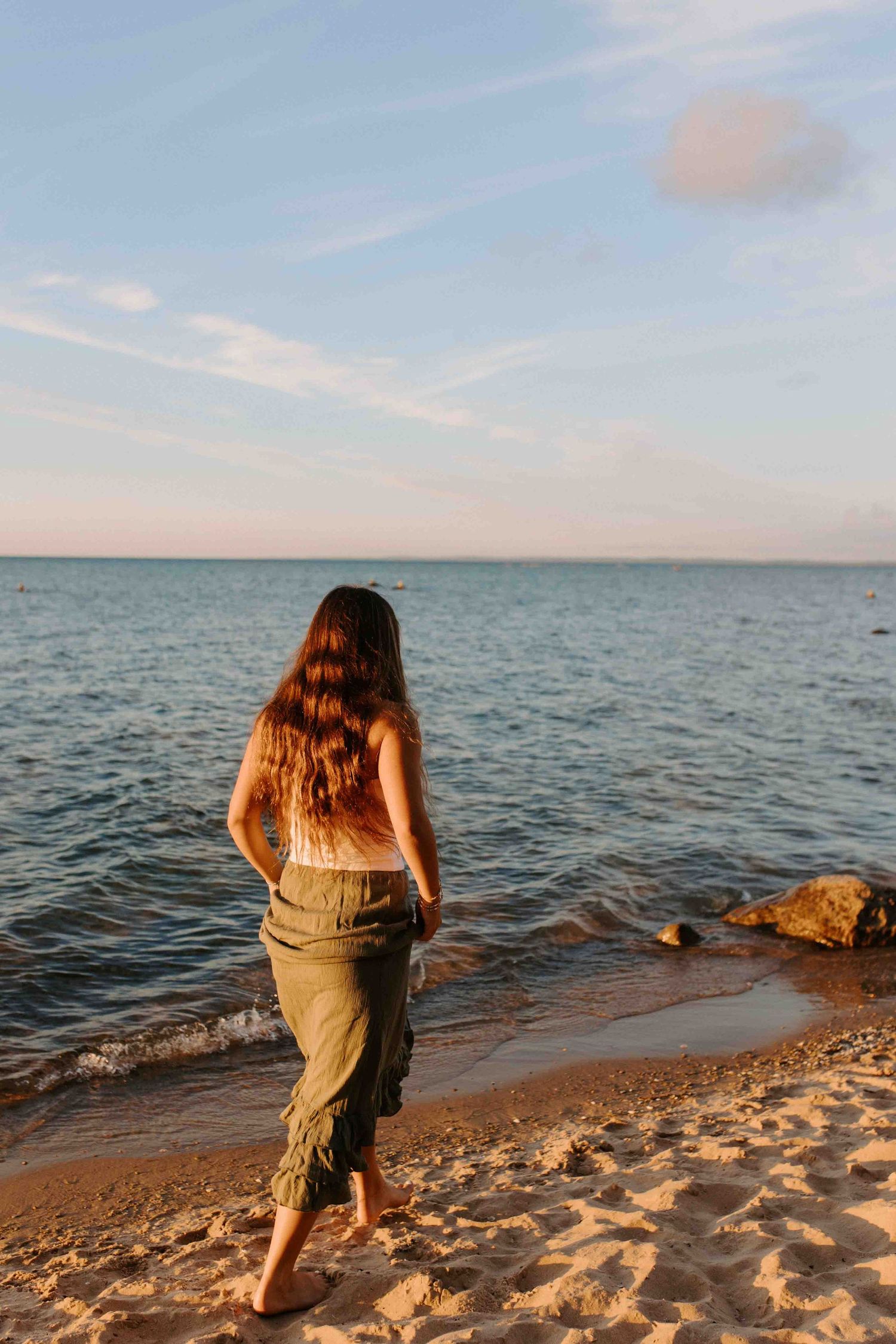 Back view of someone walking barefoot along a sandy beach at sunset with ocean waves rolling in.