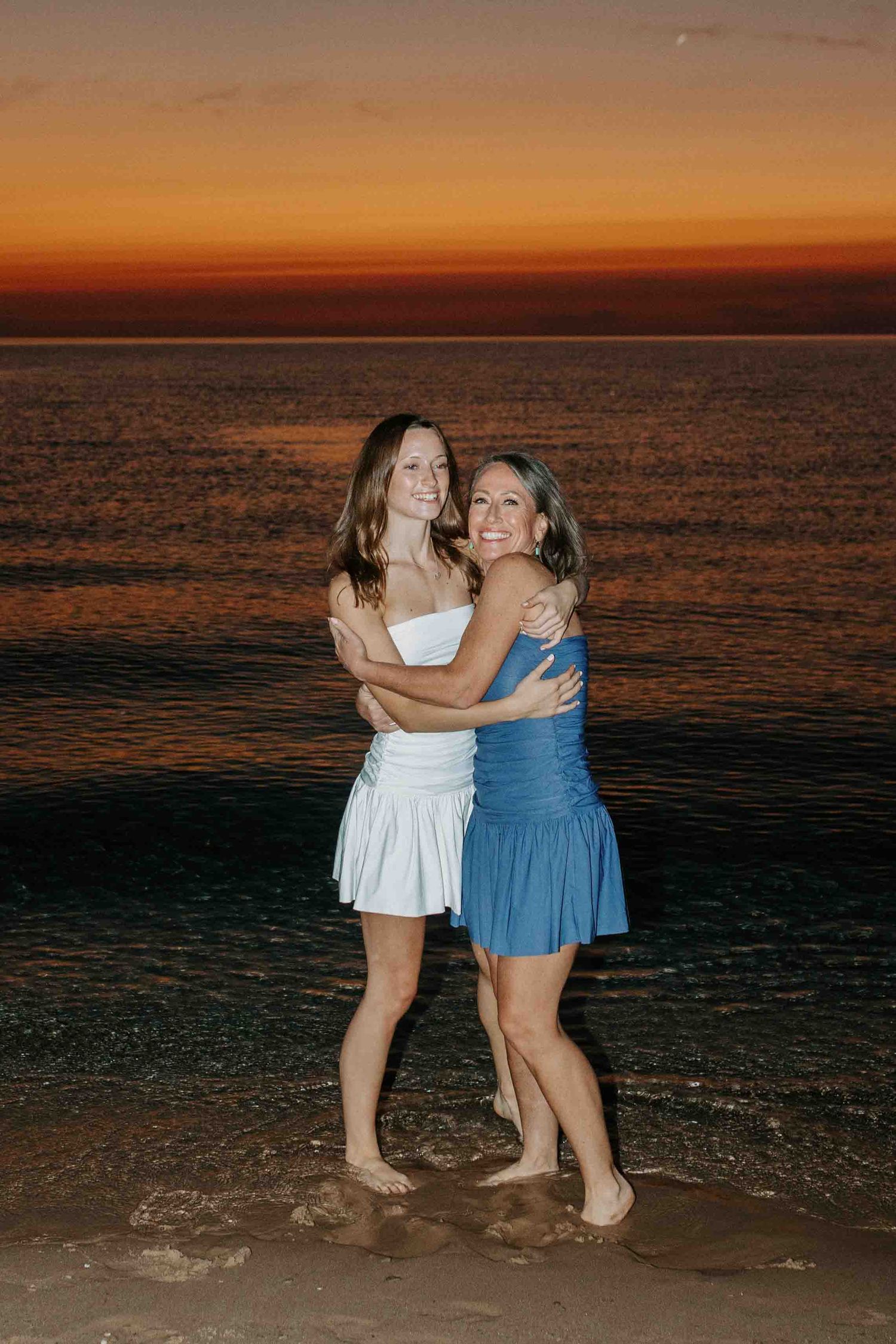 Friends in coordinating blue and white dresses hug and laugh on the beach at sunset.