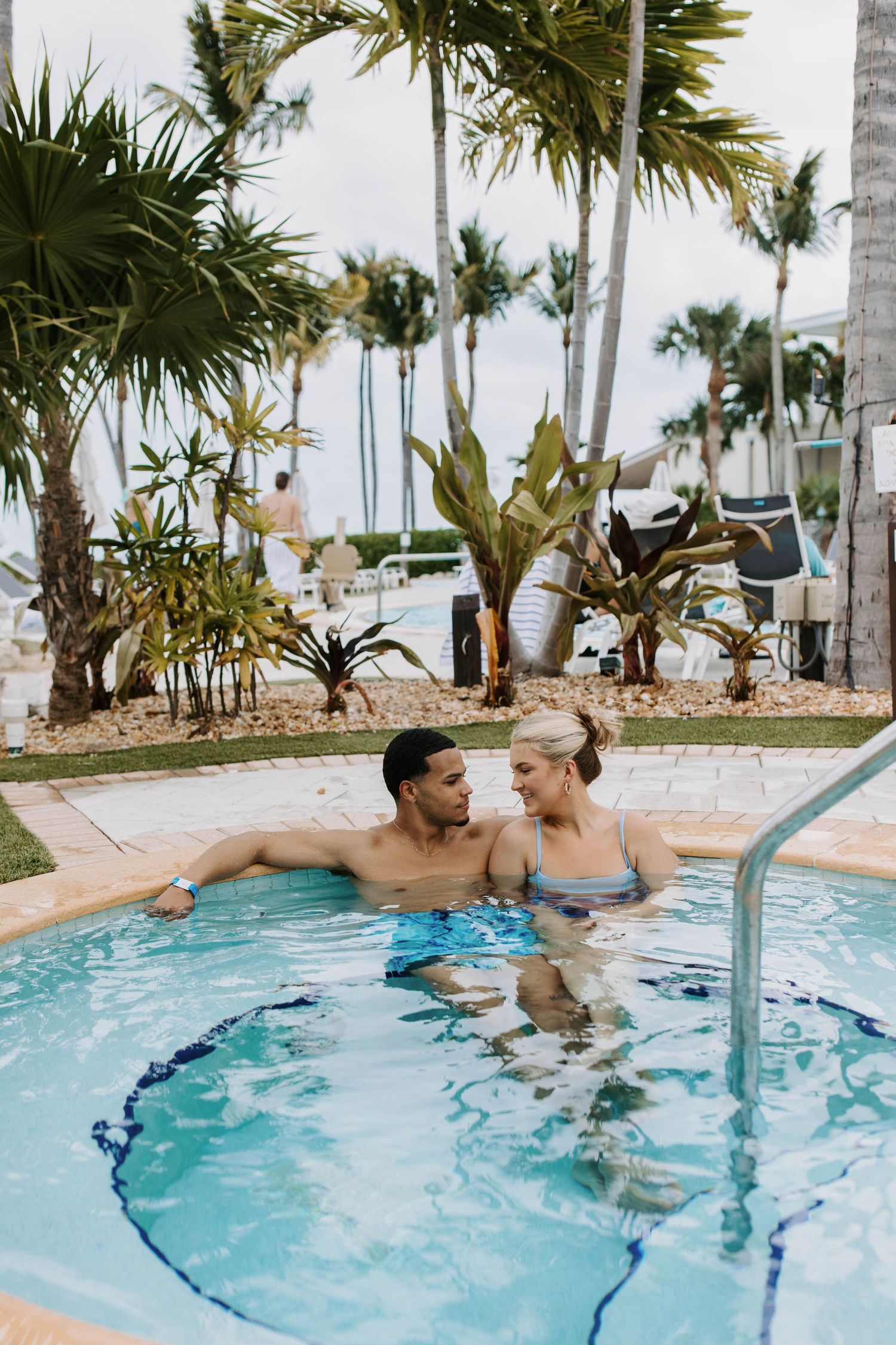 Couple relaxing in a tropical resort swimming pool surrounded by palm trees and lush landscaping.