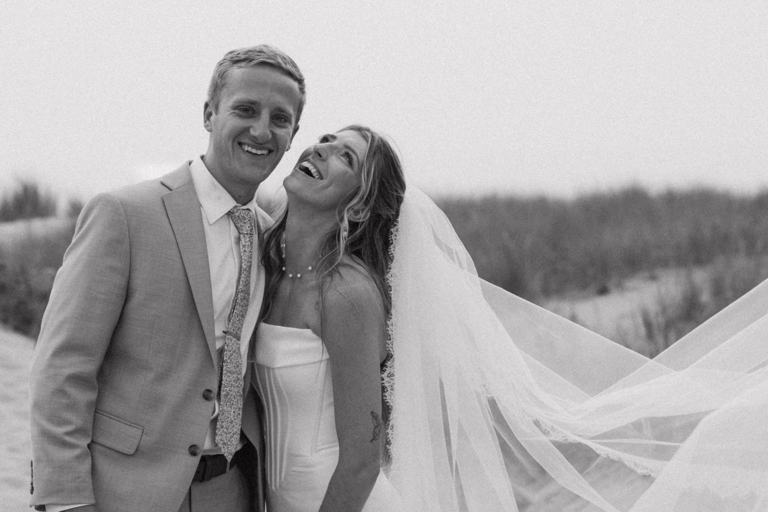 Couple in wedding attire poses together against a bright, overcast sky.
