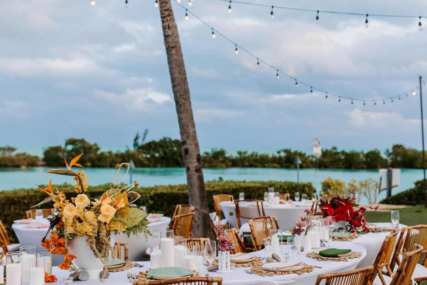 Tropical waterfront wedding reception with string lights and floral centerpieces.