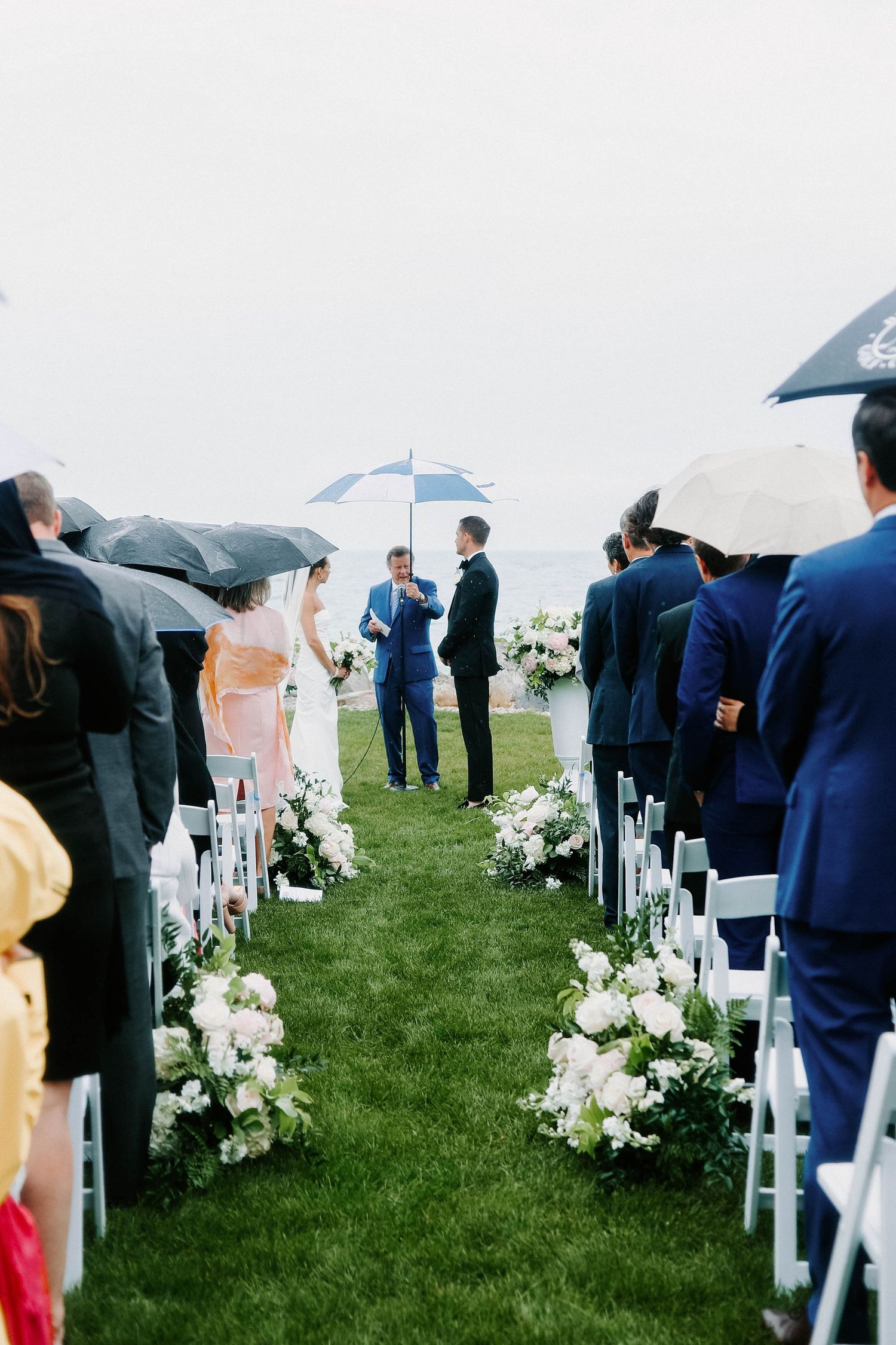 Wedding guests gather on a grassy lawn under umbrellas for an outdoor ceremony with white floral arrangements.