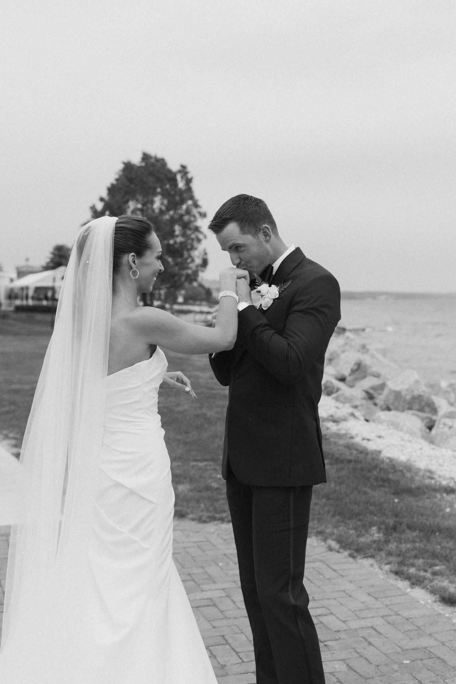 A black and white photo of a wedding couple sharing a candid moment on a brick path by the water.