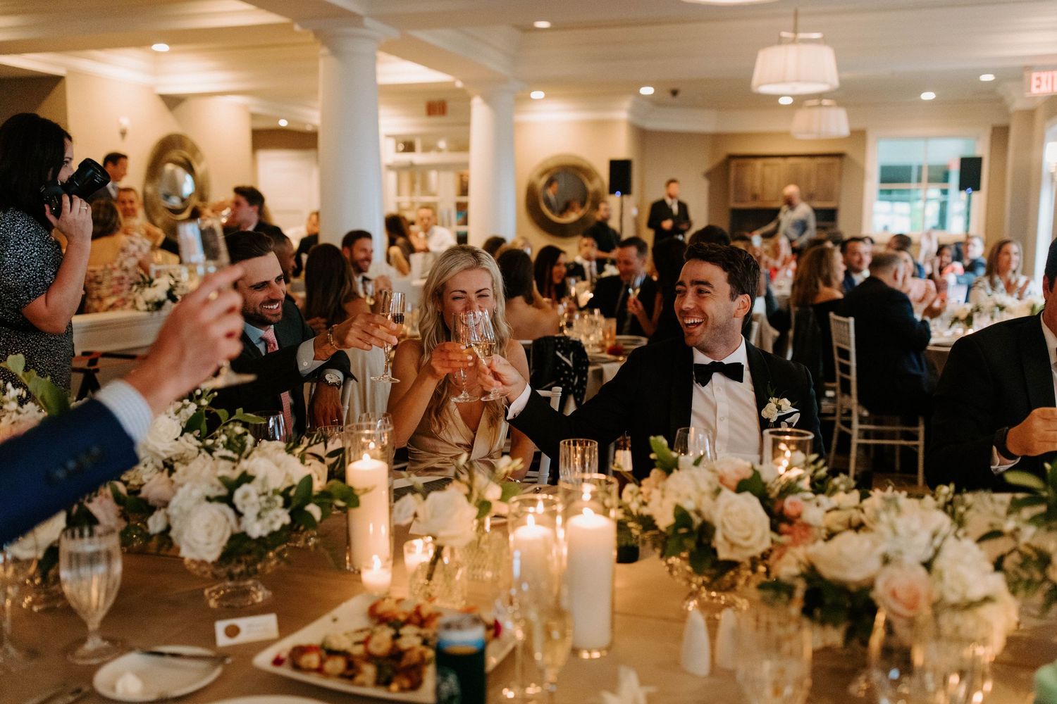 Wedding reception guests raise glasses for a toast amid candlelit tables with floral arrangements.