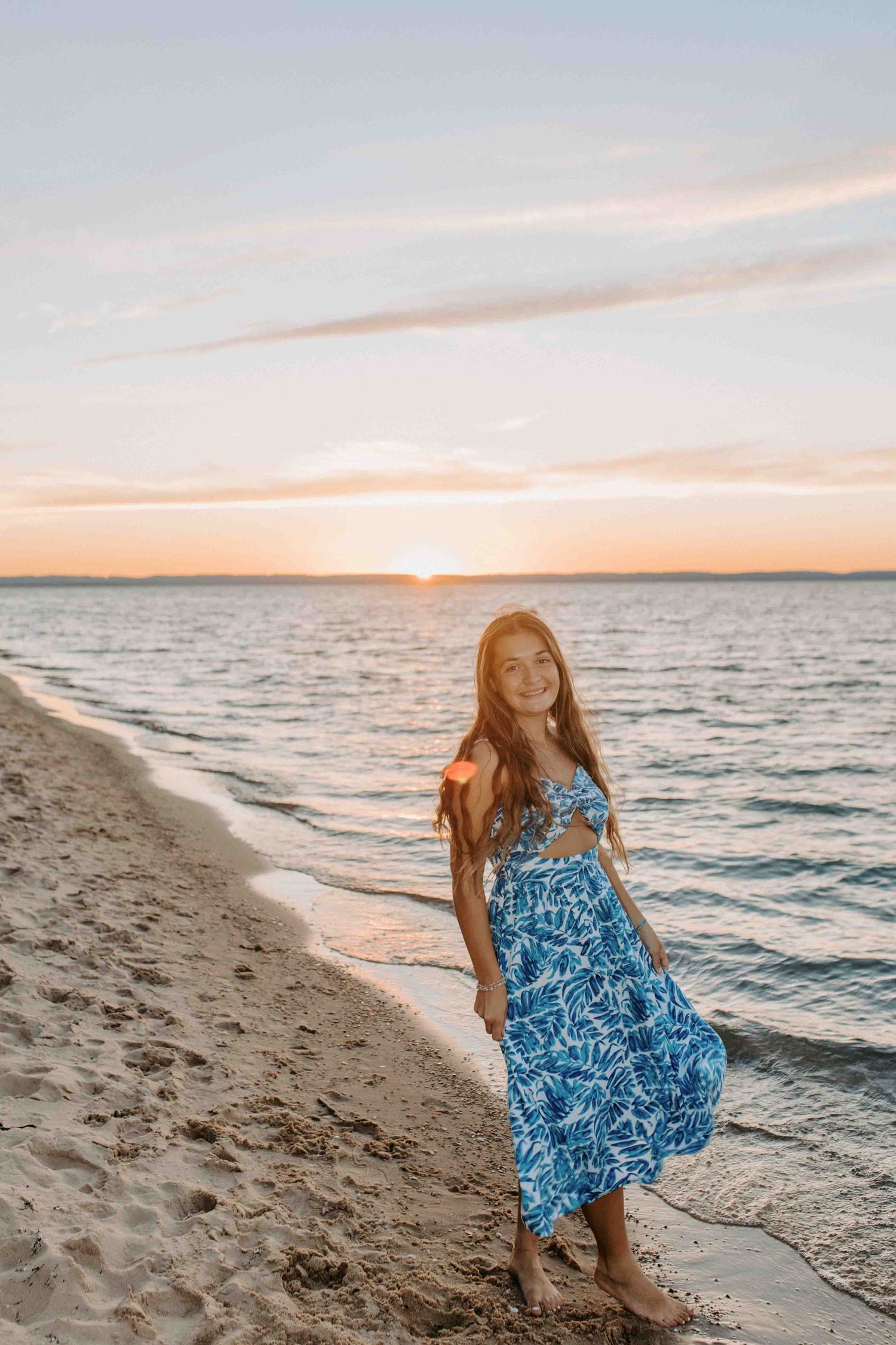 Figure in flowing blue dress poses on beach shoreline during a golden sunset.