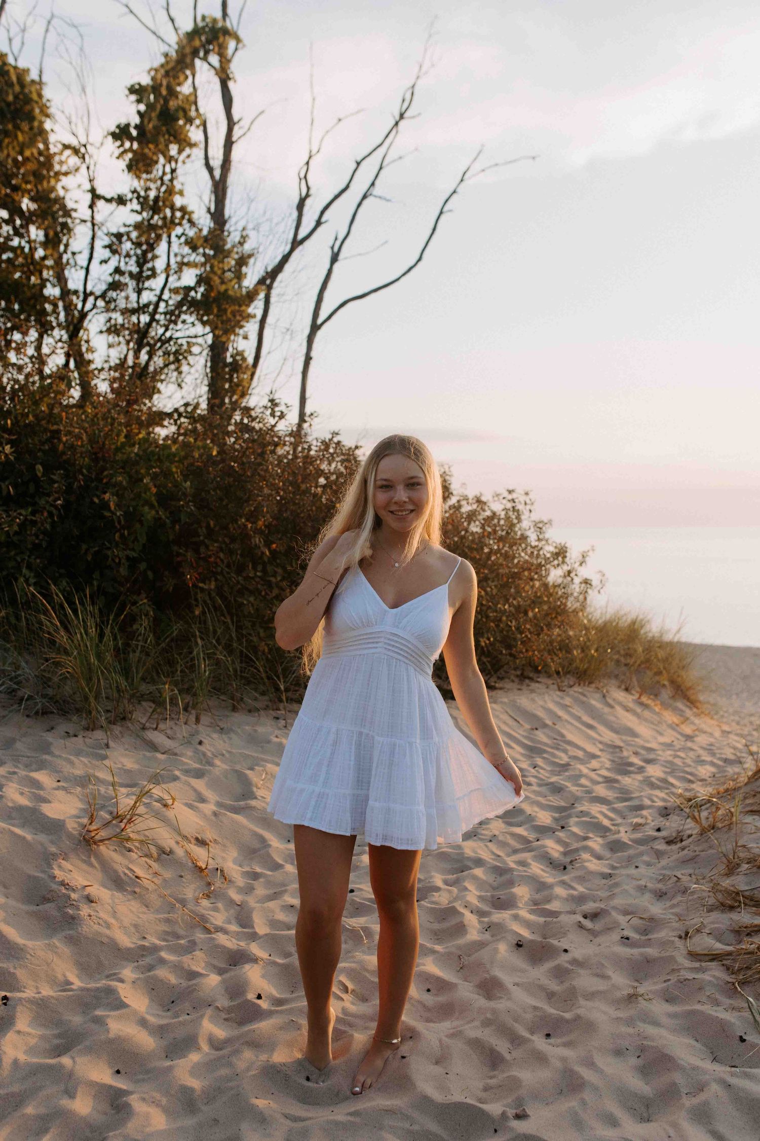 Person in white dress walking barefoot on sandy beach dunes at sunset.