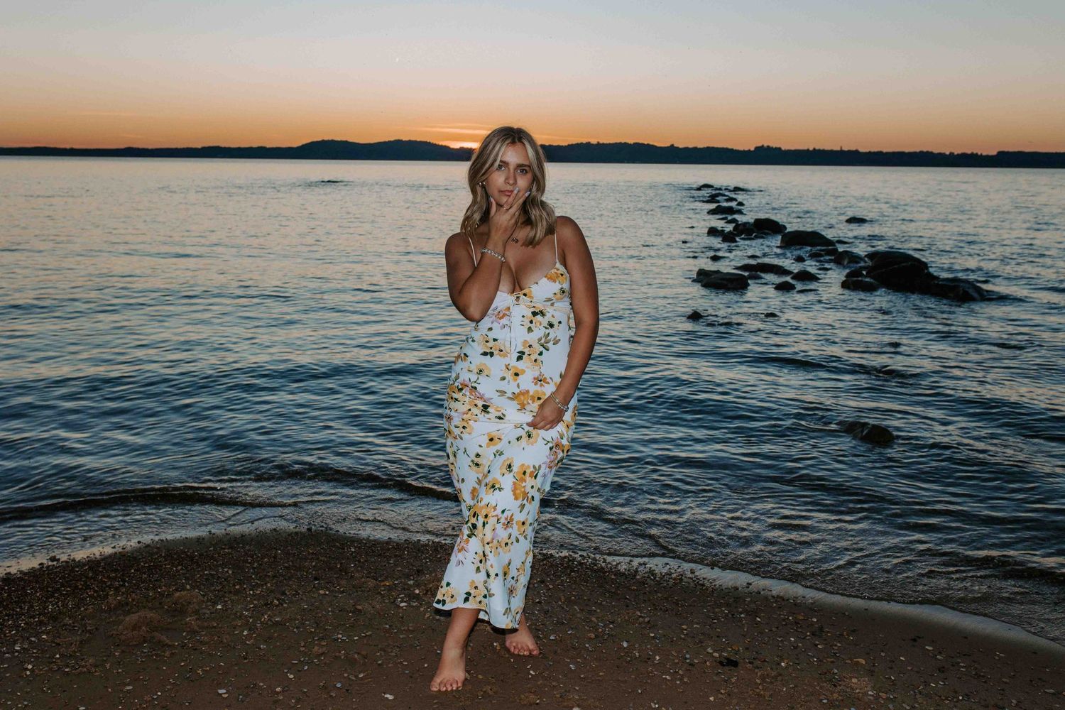 A person in a floral dress poses on a rocky beach during sunset.