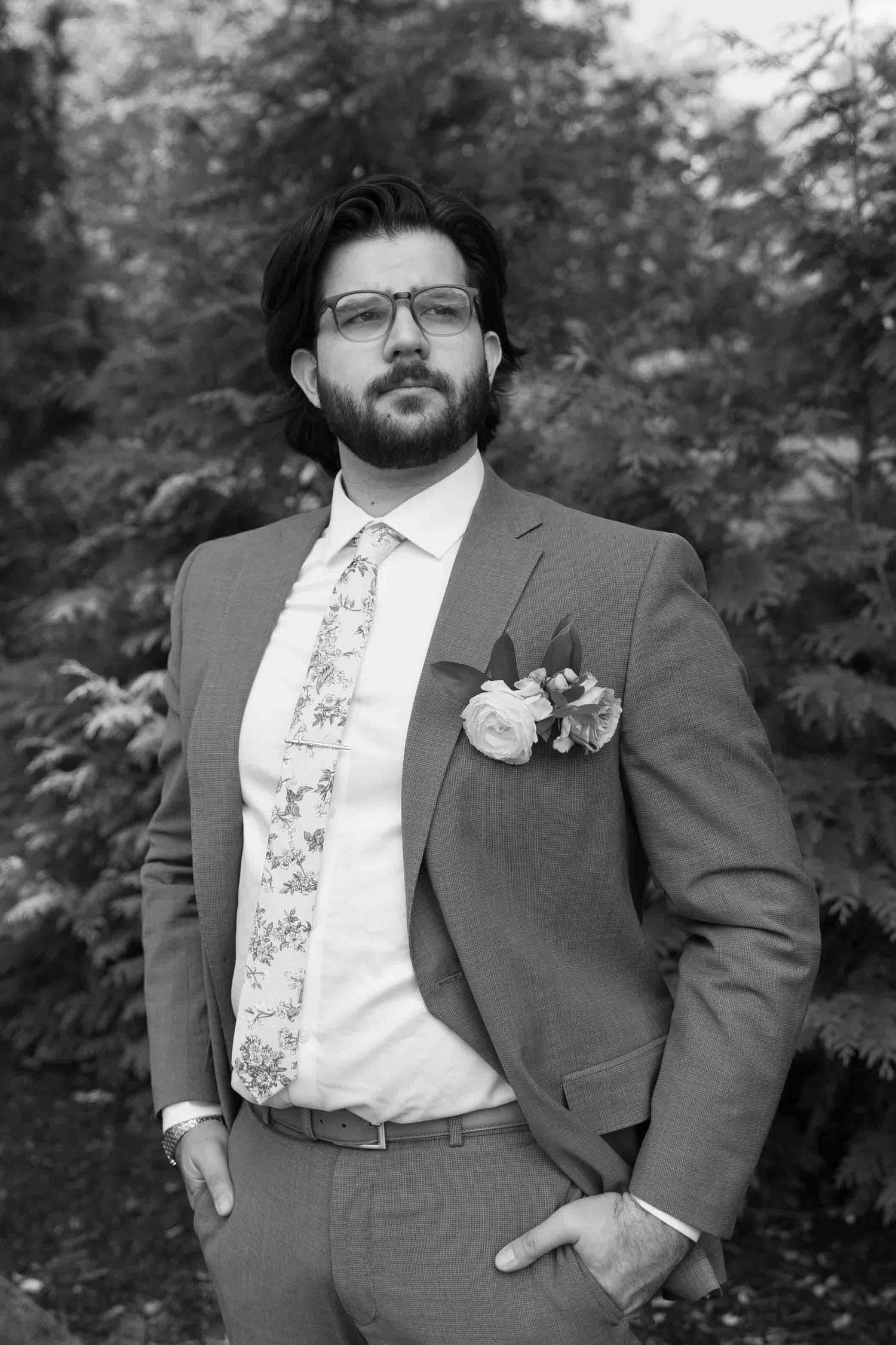 A groom in a gray suit and patterned tie poses for formal wedding portraits against a natural outdoor backdrop.