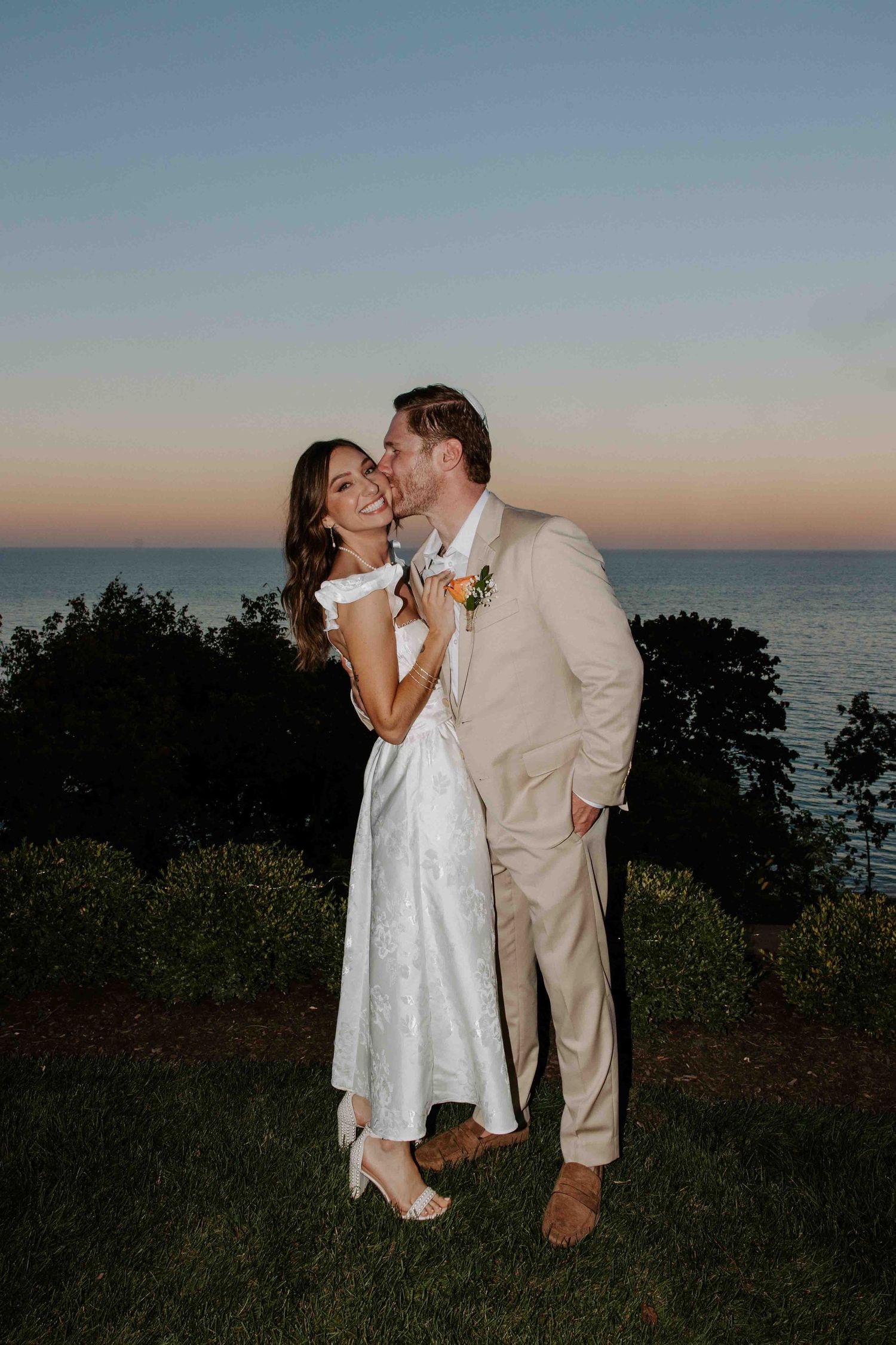 A couple shares a romantic moment against a sunset ocean backdrop in casual wedding attire.