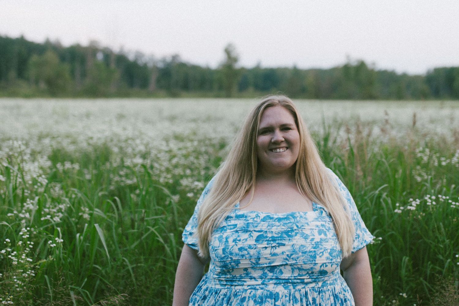 A person wearing a blue floral dress stands in a field of white wildflowers on a sunny day.