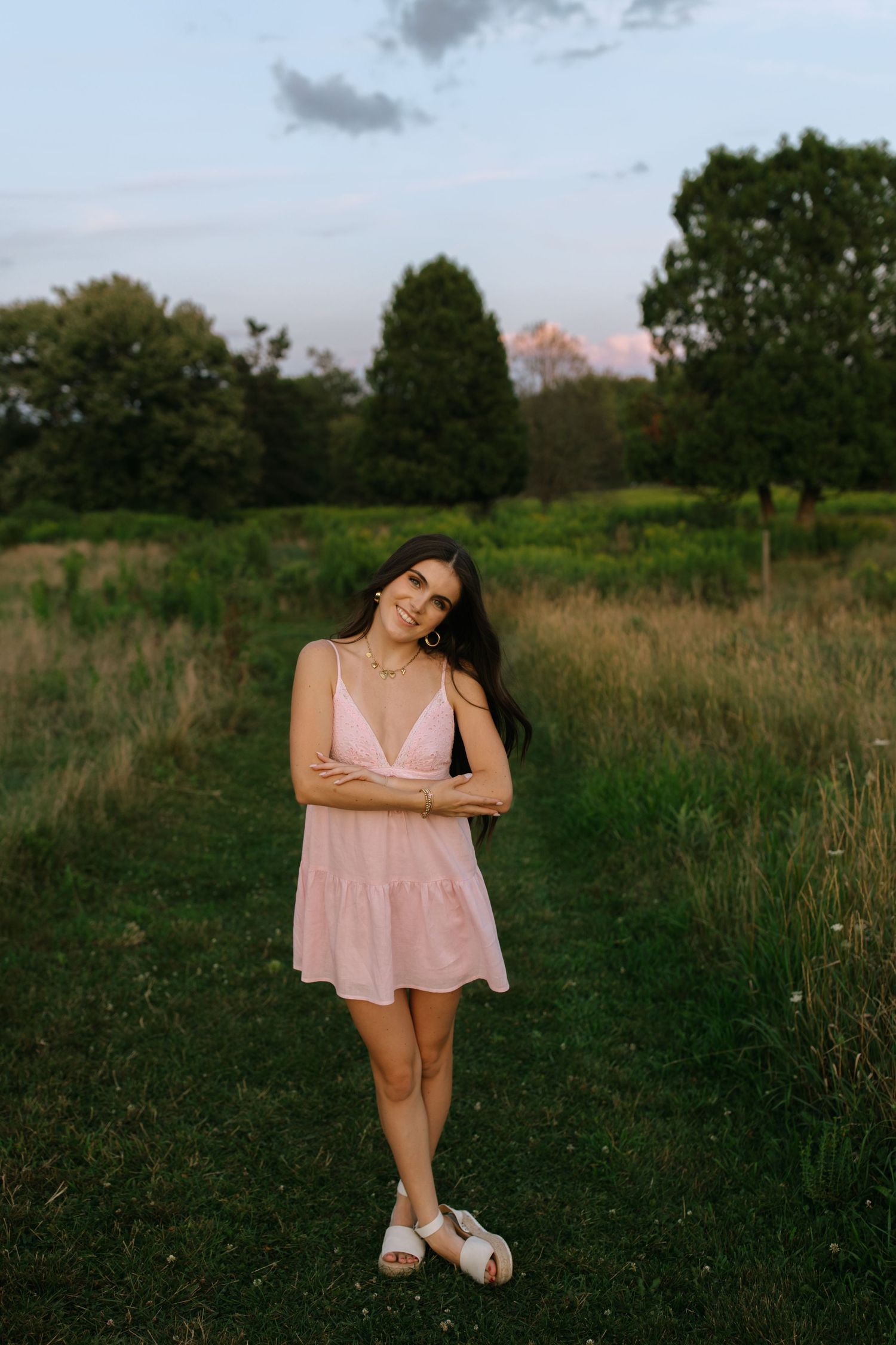Woman in pink dress poses casually in a rustic field at sunset.