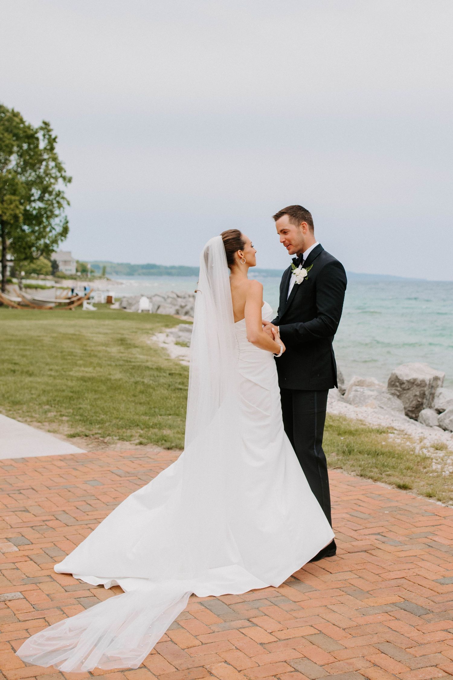 A couple in wedding attire poses on a brick patio overlooking a scenic waterfront.
