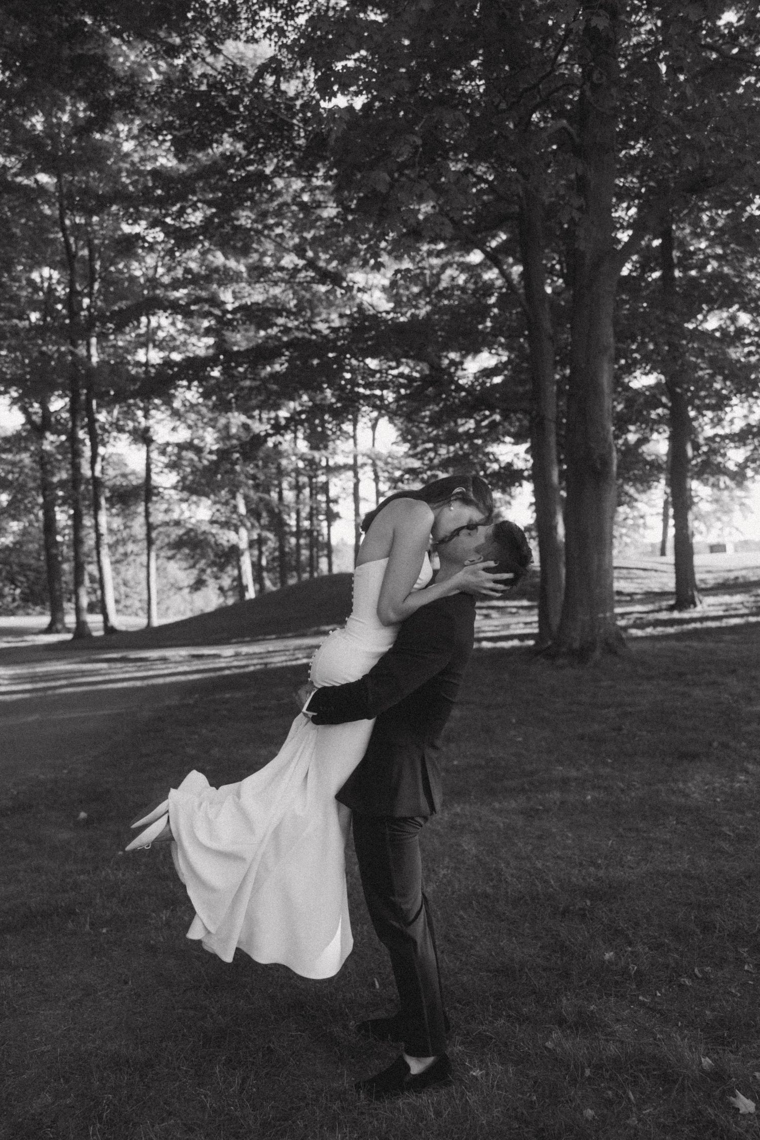 Black and white wedding portrait of couple dancing on grassy hill among trees.