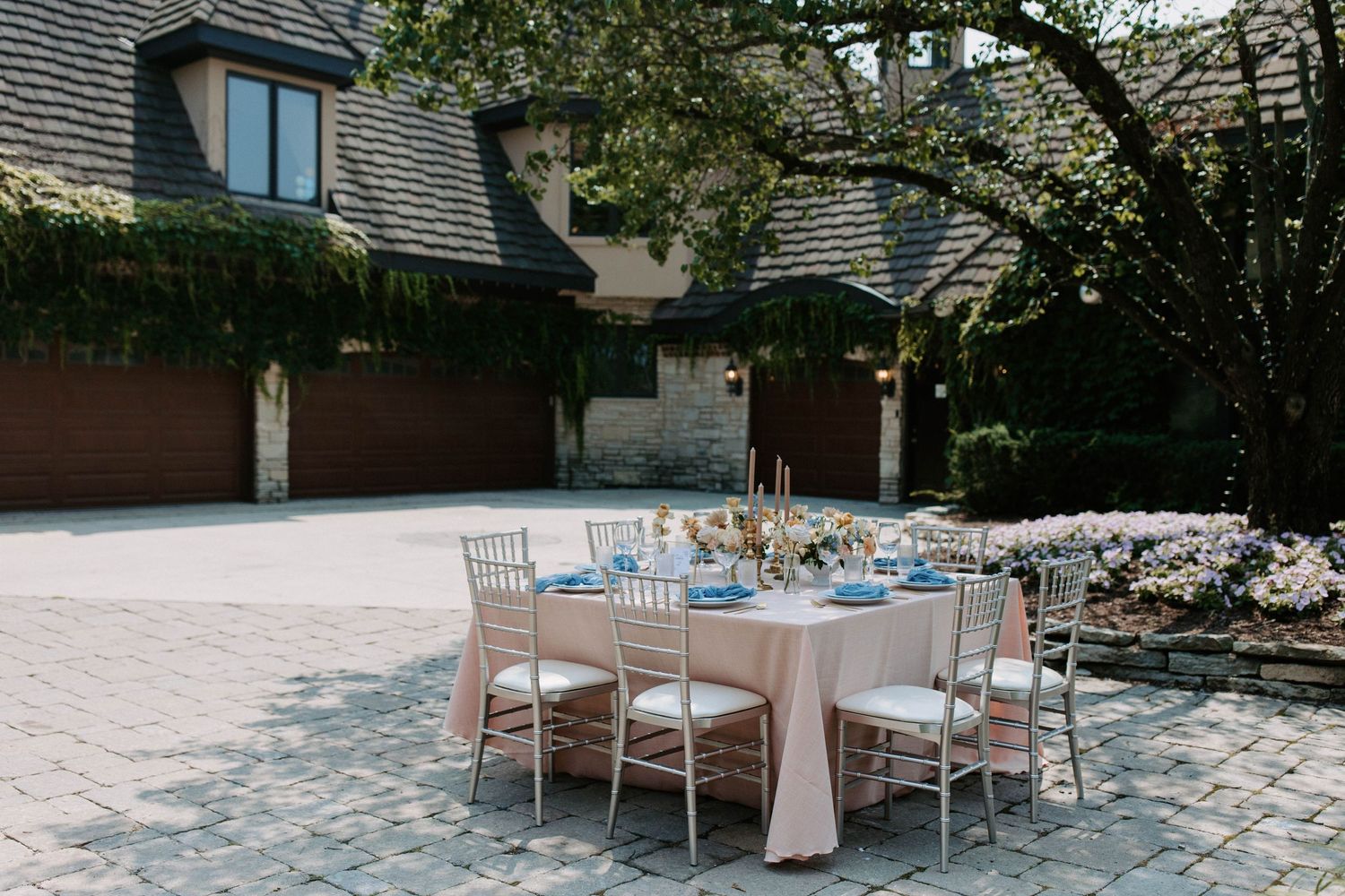 Elegant outdoor wedding reception setup with white chairs and floral arrangements on stone patio surrounded by greenery.