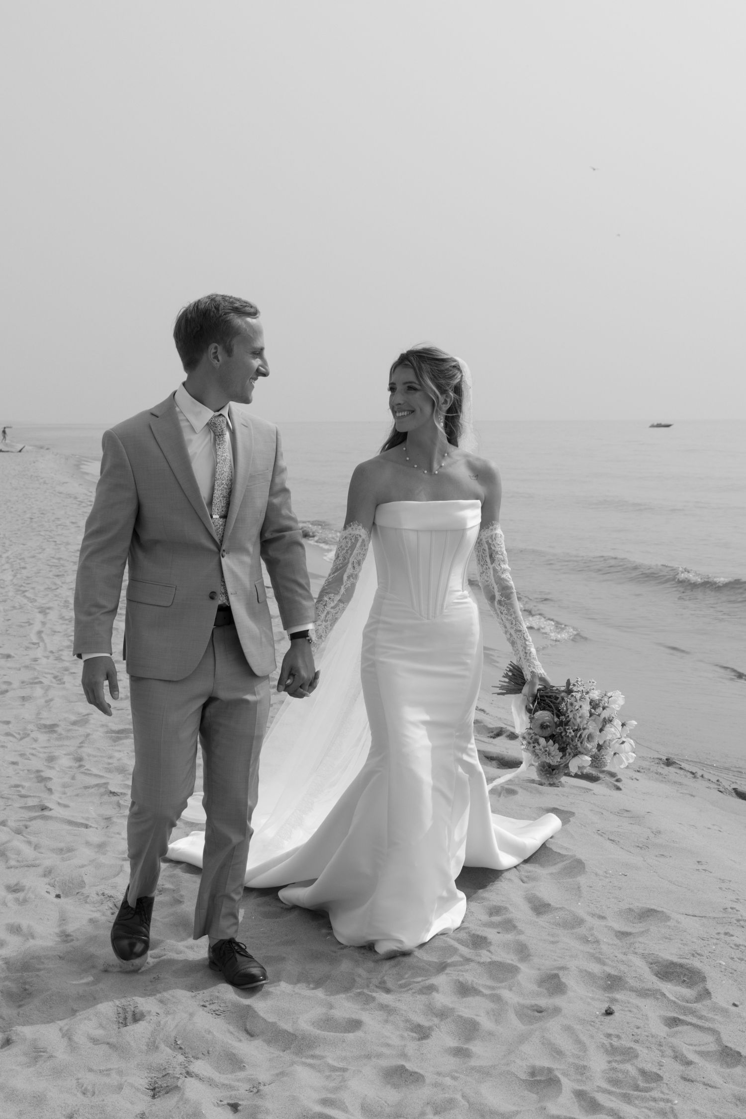 A couple walks hand in hand along a sandy beach during their wedding photoshoot.