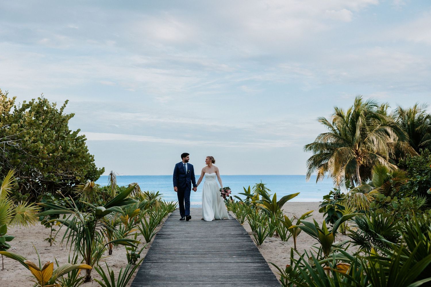 A couple in wedding attire walks along a wooden boardwalk surrounded by tropical scenery.