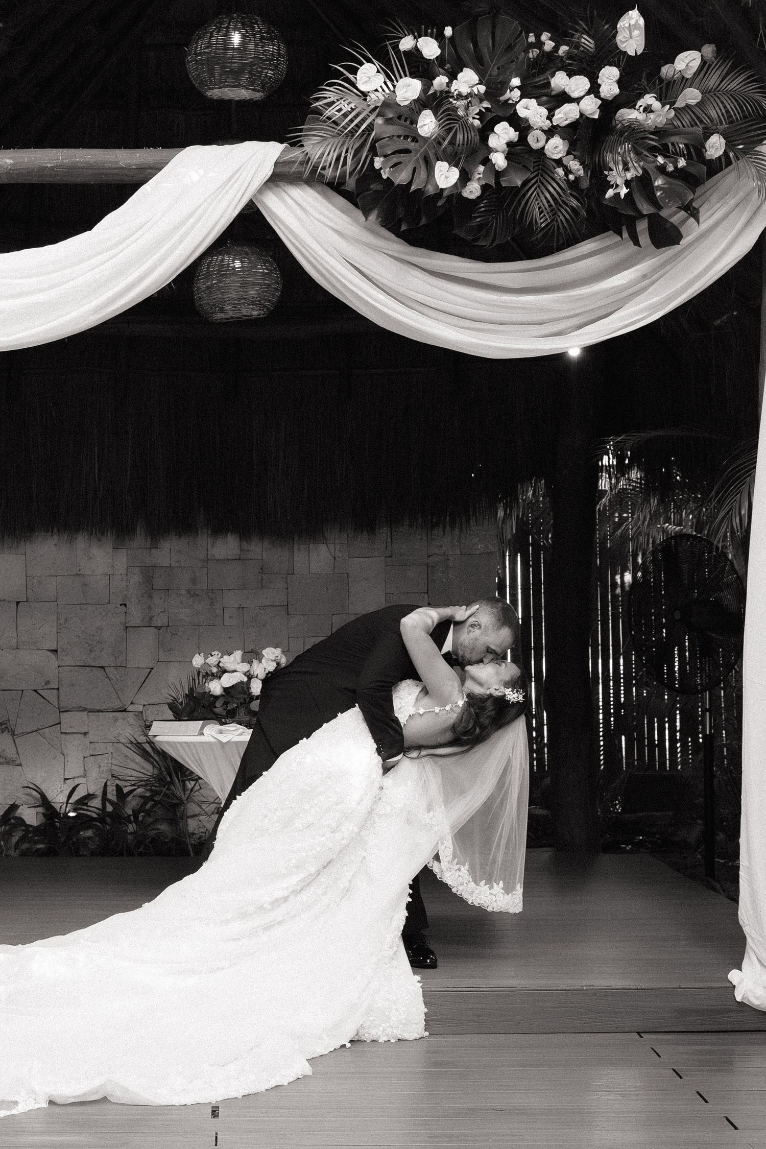 A romantic dip kiss under draped fabric and floral decorations at a wedding venue.